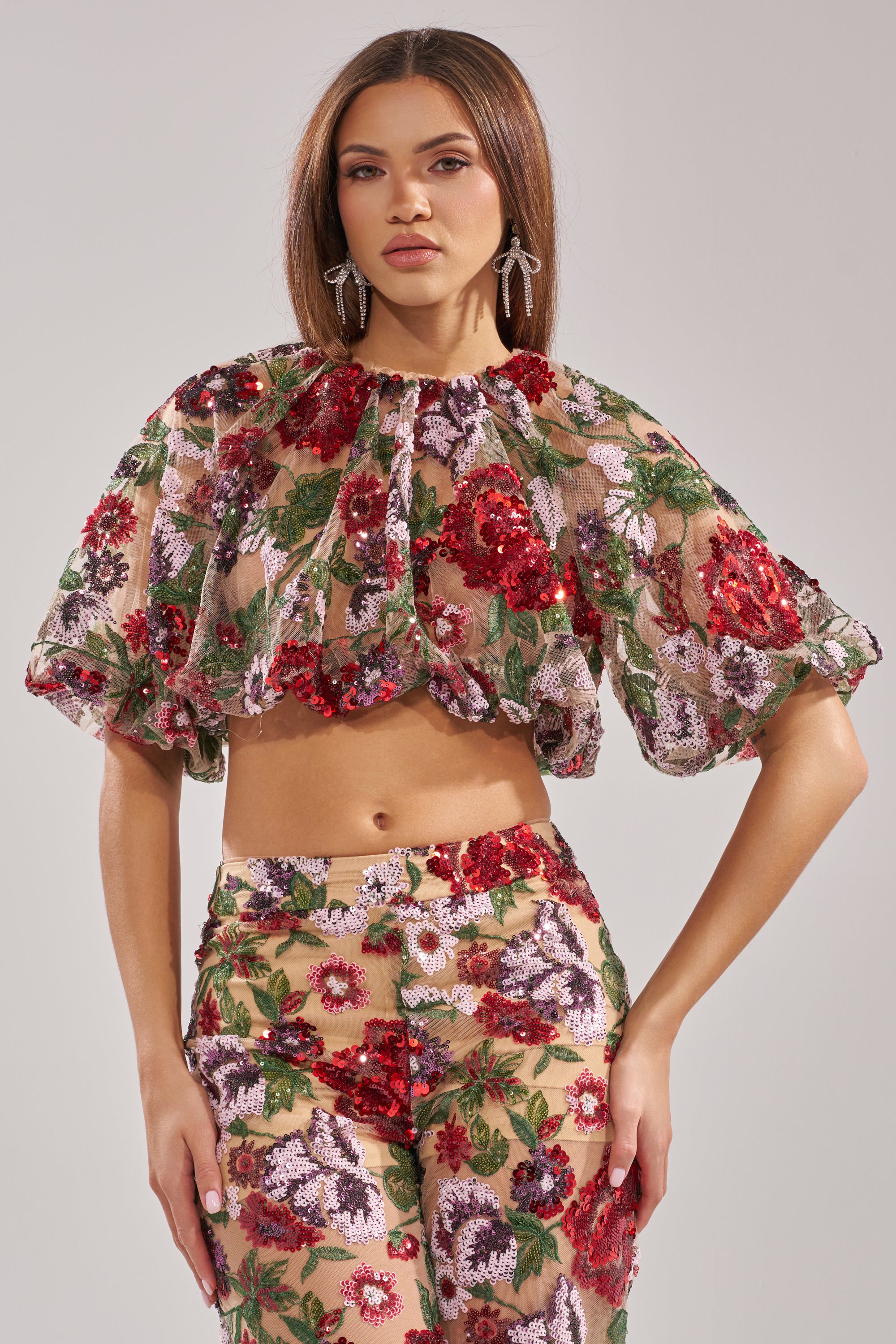 A woman models the ENCHANTED GARDEN FLORAL EMBROIDERED TULLE TOP, featuring puffed sleeves and vivid red, green, and purple floral patterns on sheer tulle, styled with matching high-waisted pants against a plain background.