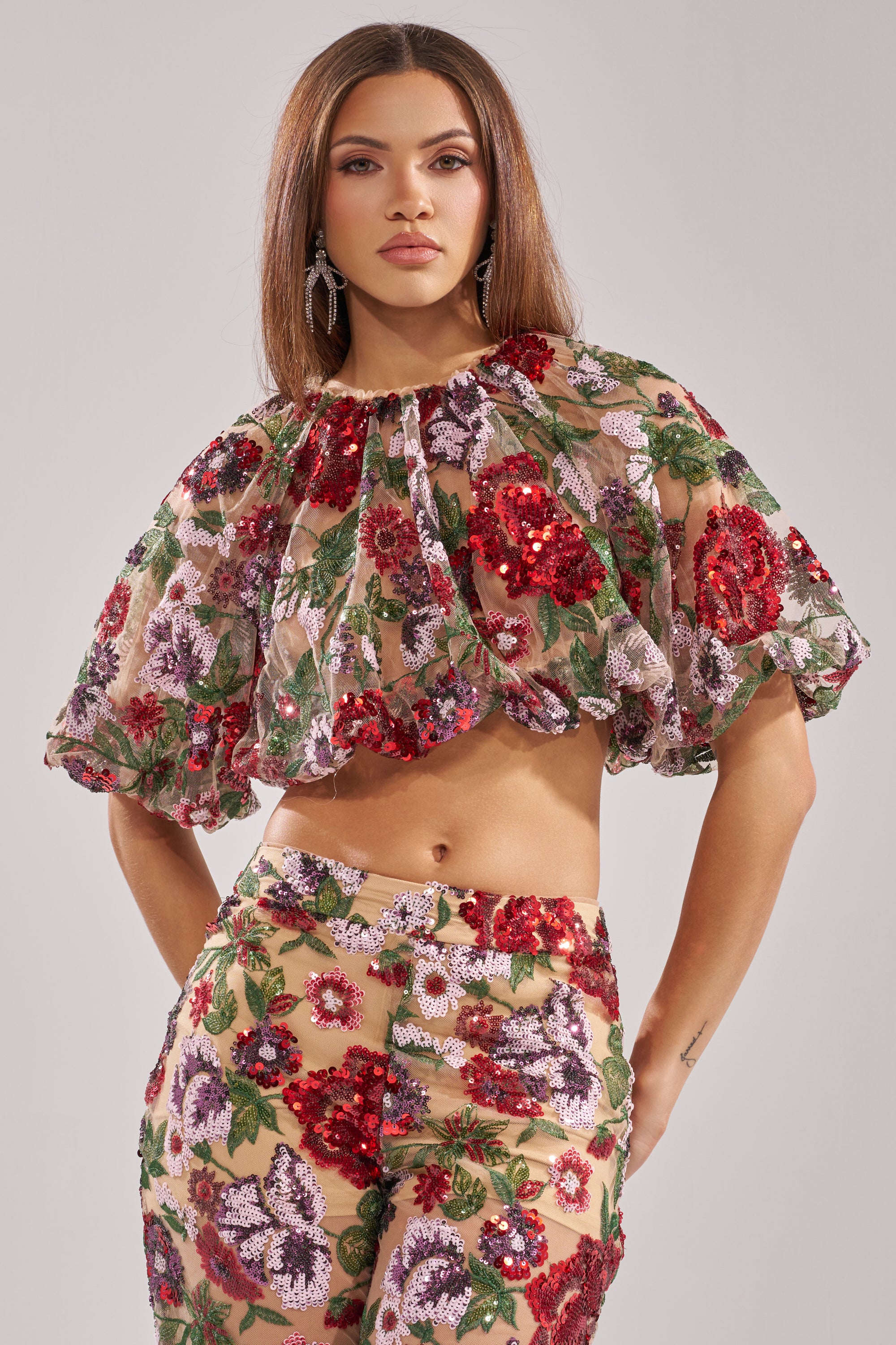 A woman with straight brown hair and statement earrings poses confidently in the ENCHANTED GARDEN FLORAL EMBROIDERED TULLE TOP—a flared crop of sheer tulle adorned with red, pink, and purple floral sequins—against a plain background.