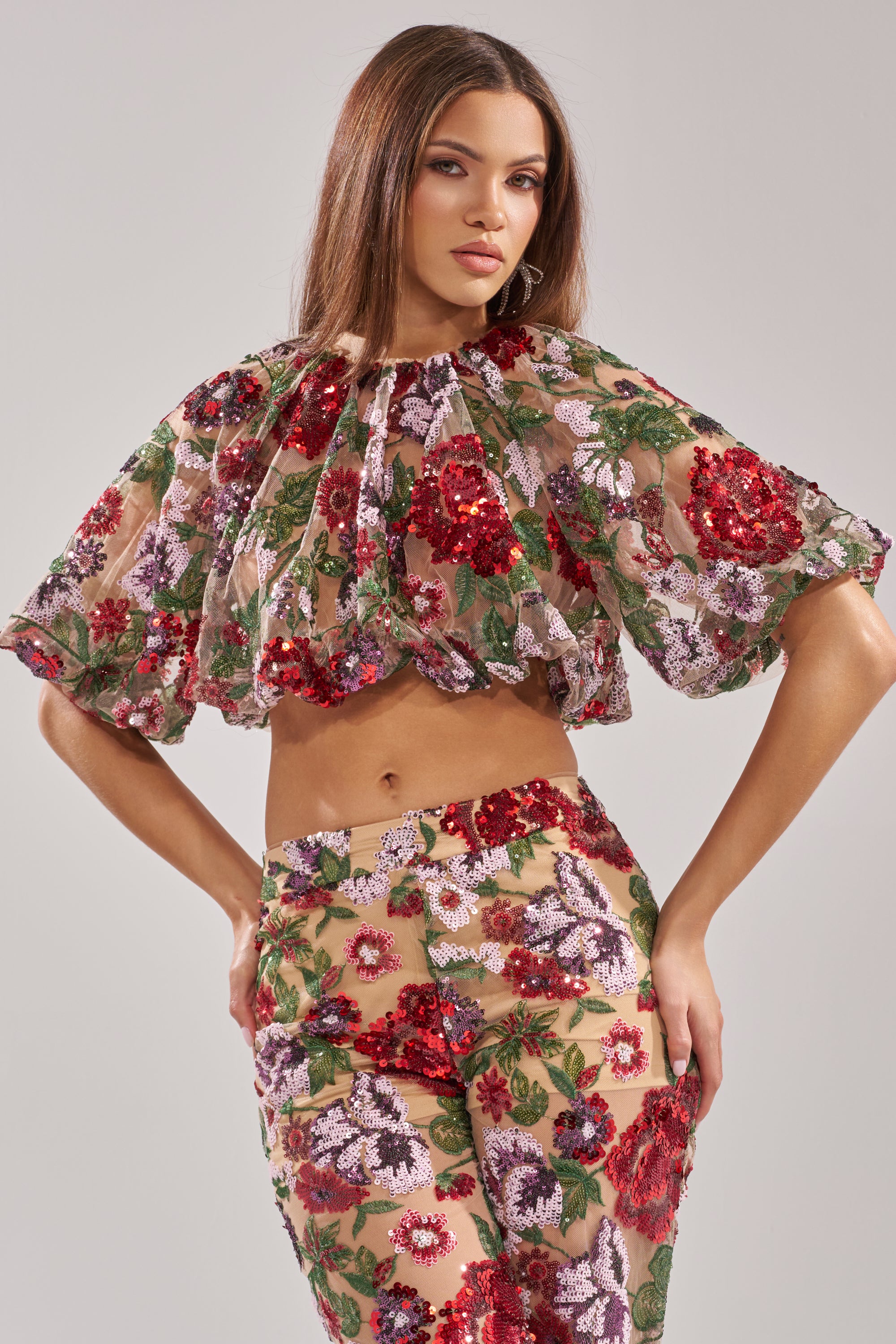 A woman stands confidently against a light gray background, wearing the ENCHANTED GARDEN FLORAL EMBROIDERED TULLE TOP—sheer with ruffled sleeves—and matching high-waisted pants adorned with red, pink, and green sequins.