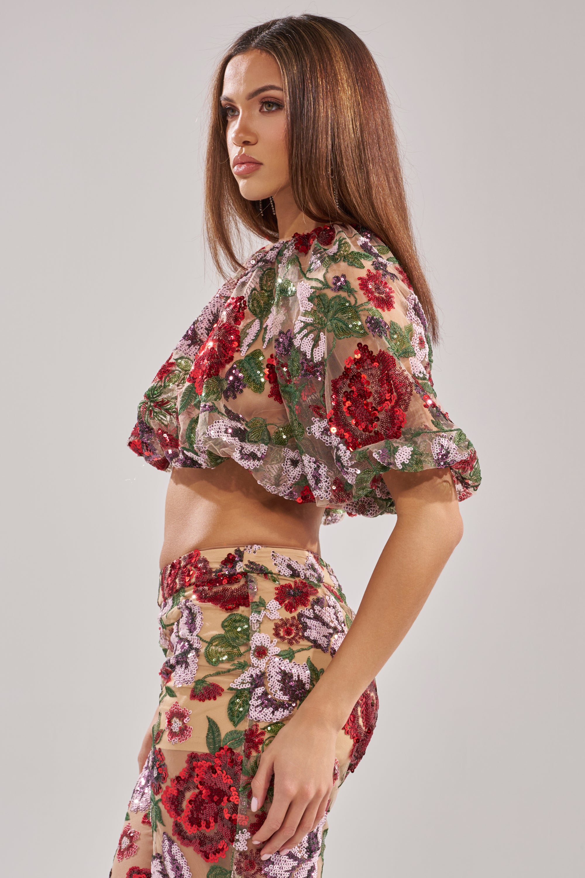 A woman with straight brown hair stands sideways against a light gray background, modeling the ENCHANTED GARDEN FLORAL EMBROIDERED TULLE TOP, featuring sheer fabric adorned with red, pink, and green floral embroidery.