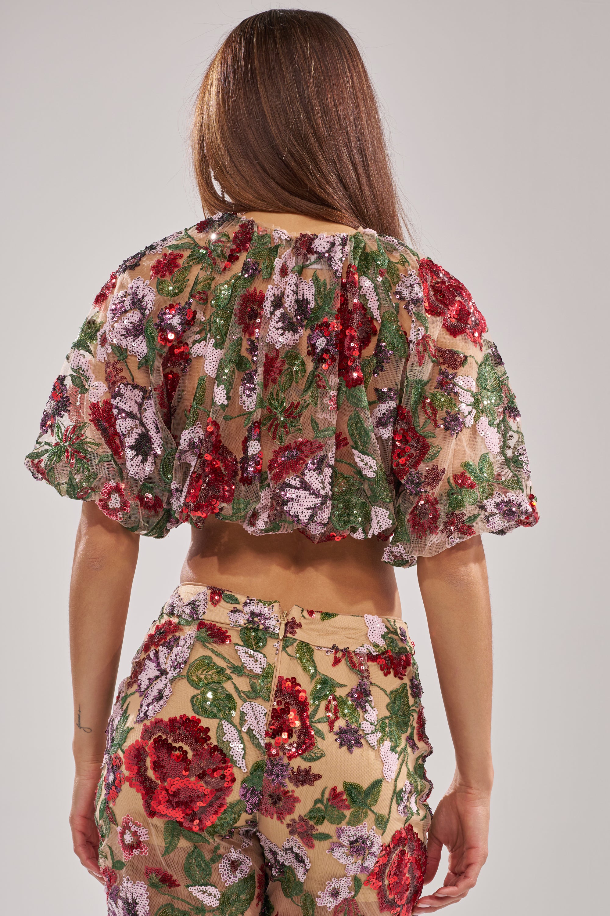 A woman, seen from behind with her brown hair down, wears the ENCHANTED GARDEN FLORAL EMBROIDERED TULLE TOP. The sheer blouse features red, purple, and green floral embroidery and sequins, paired with matching high-waisted pants.