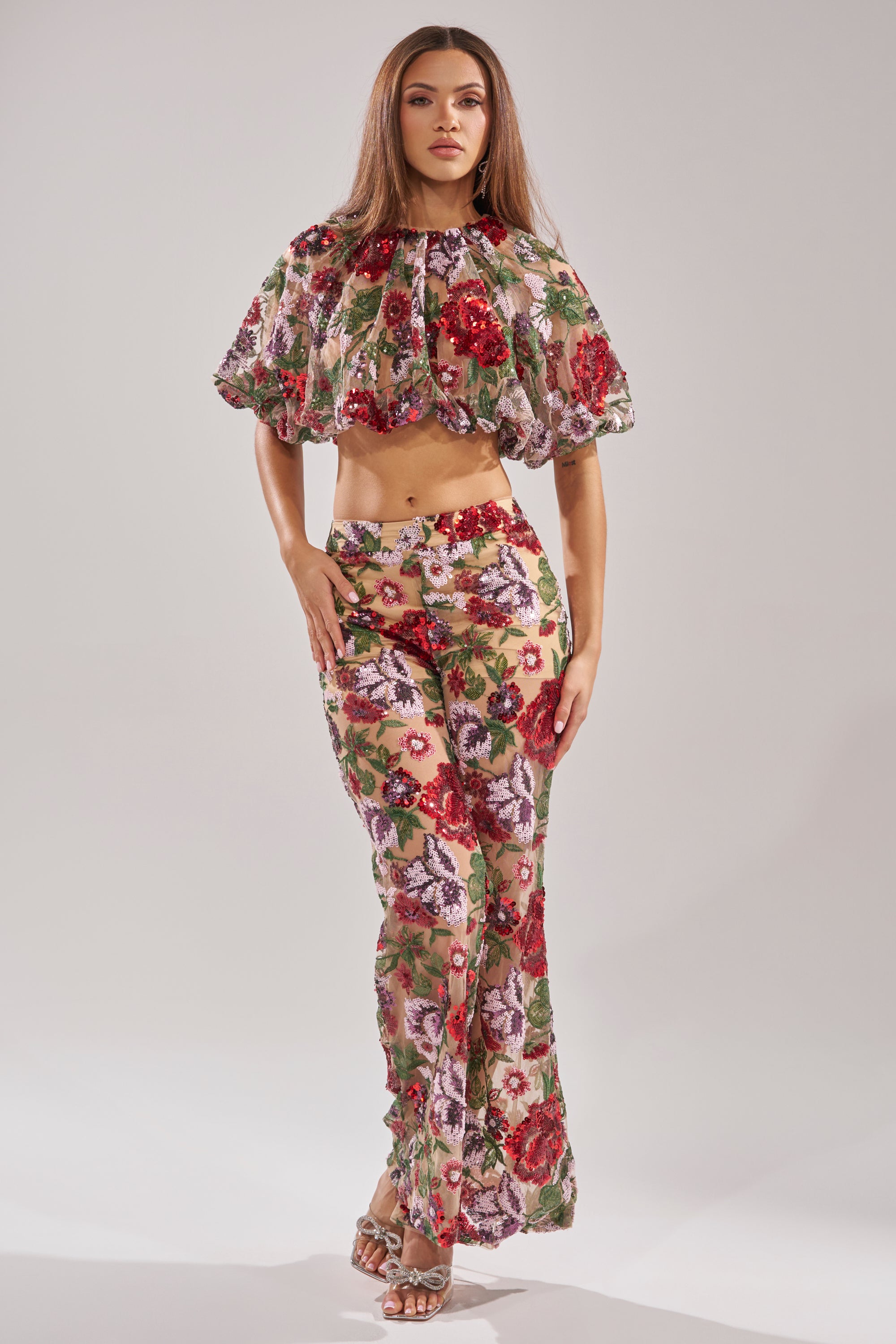 A woman stands confidently in the ENCHANTED GARDEN FLORAL EMBROIDERED TULLE TOP with puffed sleeves and matching high-waisted pants. She has long straight hair, open-toe heels, and poses against a plain light background.