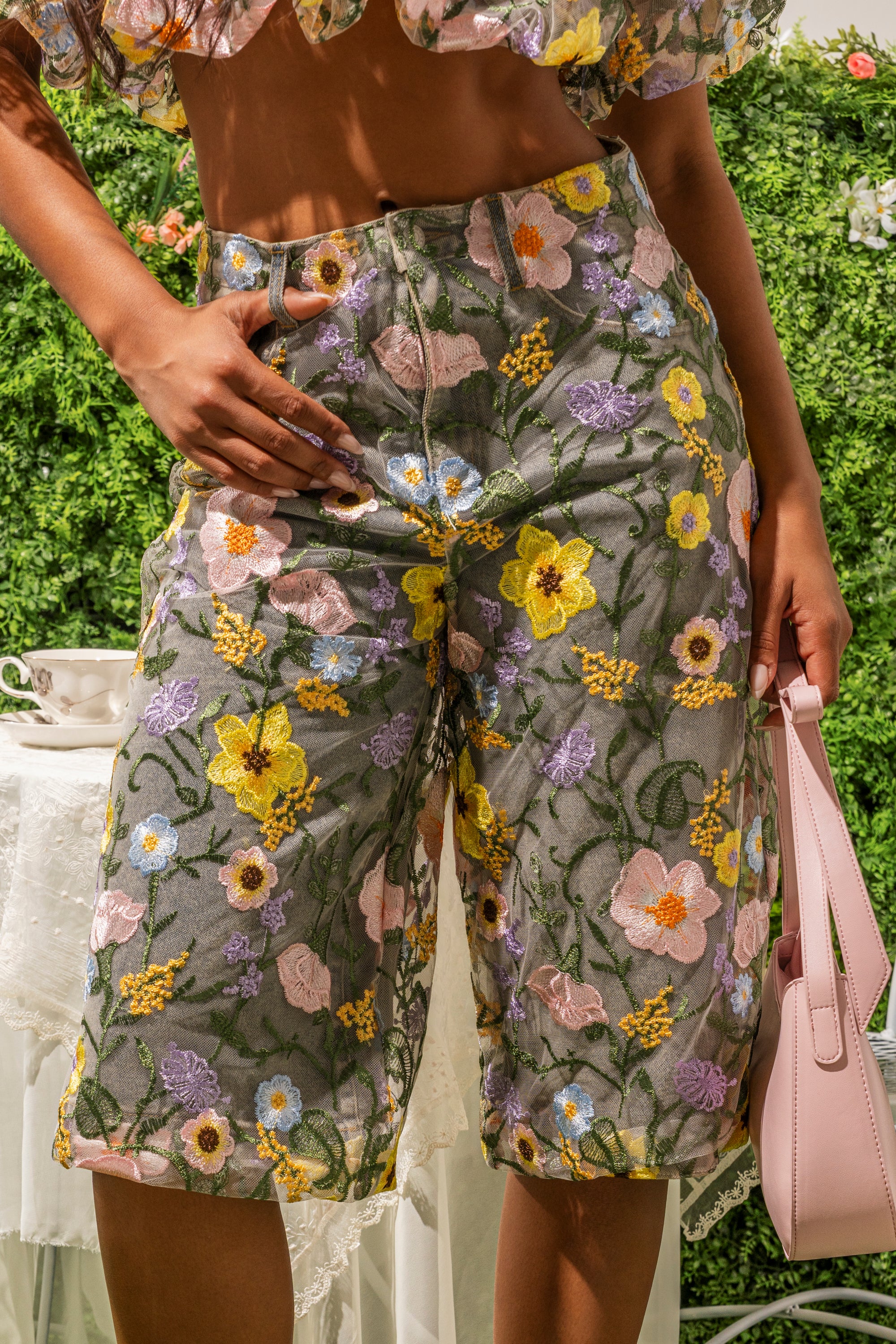 Outdoors, someone holds a light pink handbag while wearing GARDEN PARTY STRETCH DENIM WITH FLORAL TULLE BERMUDA SHORTS. Greenery and a white lace-covered table appear in the background. 0