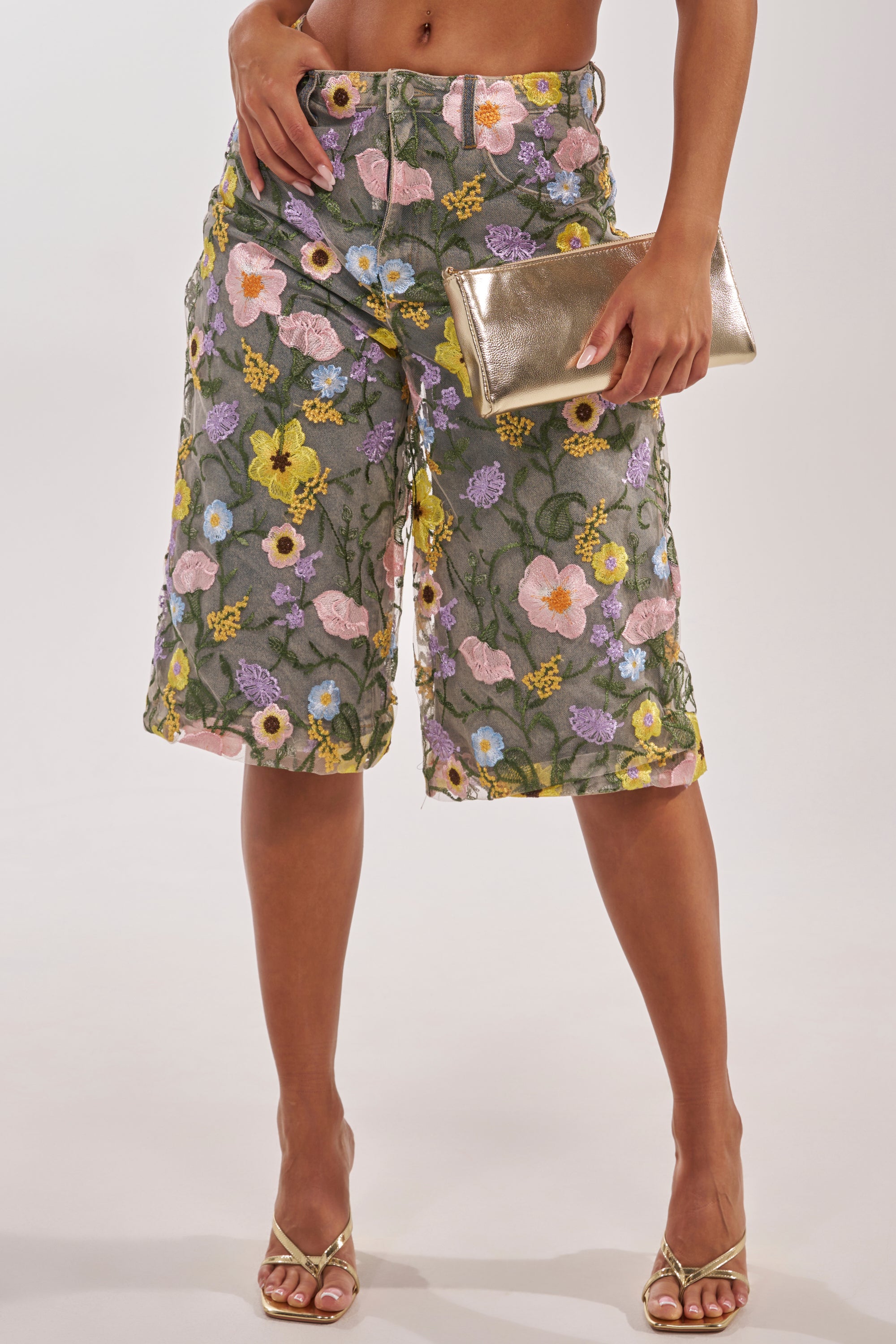 A person wearing GARDEN PARTY STRETCH DENIM WITH FLORAL TULLE BERMUDA SHORTS featuring pastel floral embroidery, gold heeled sandals, and holding a metallic gold clutch against a plain light background. 3