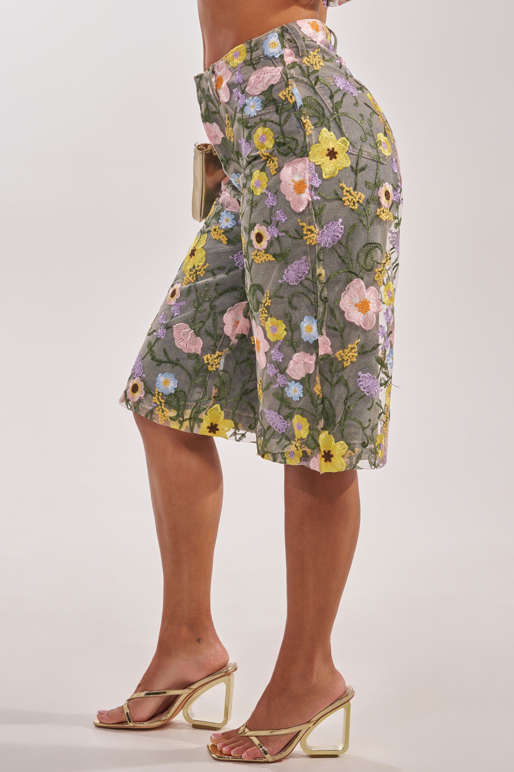 A person wears the GARDEN PARTY STRETCH DENIM WITH FLORAL TULLE BERMUDA SHORTS, featuring sheer gray fabric with colorful floral embroidery, paired with gold high-heeled sandals and a beige clutch against a plain light background. 5