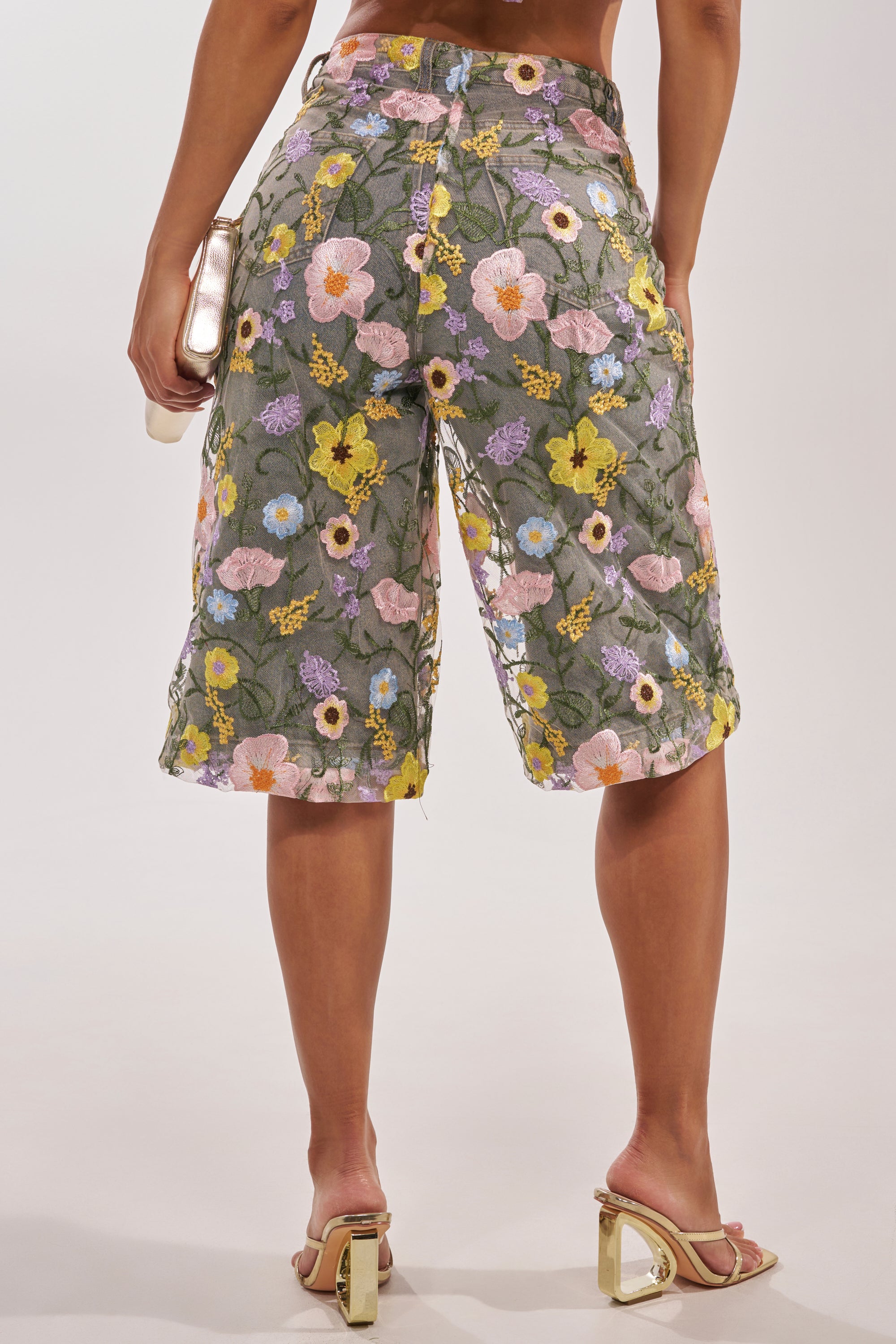 A person is seen from behind wearing GARDEN PARTY STRETCH DENIM WITH FLORAL TULLE BERMUDA SHORTS, a clutch in hand and gold high-heeled sandals, featuring knee-length, high-rise fit with a green floral tulle overlay. 6