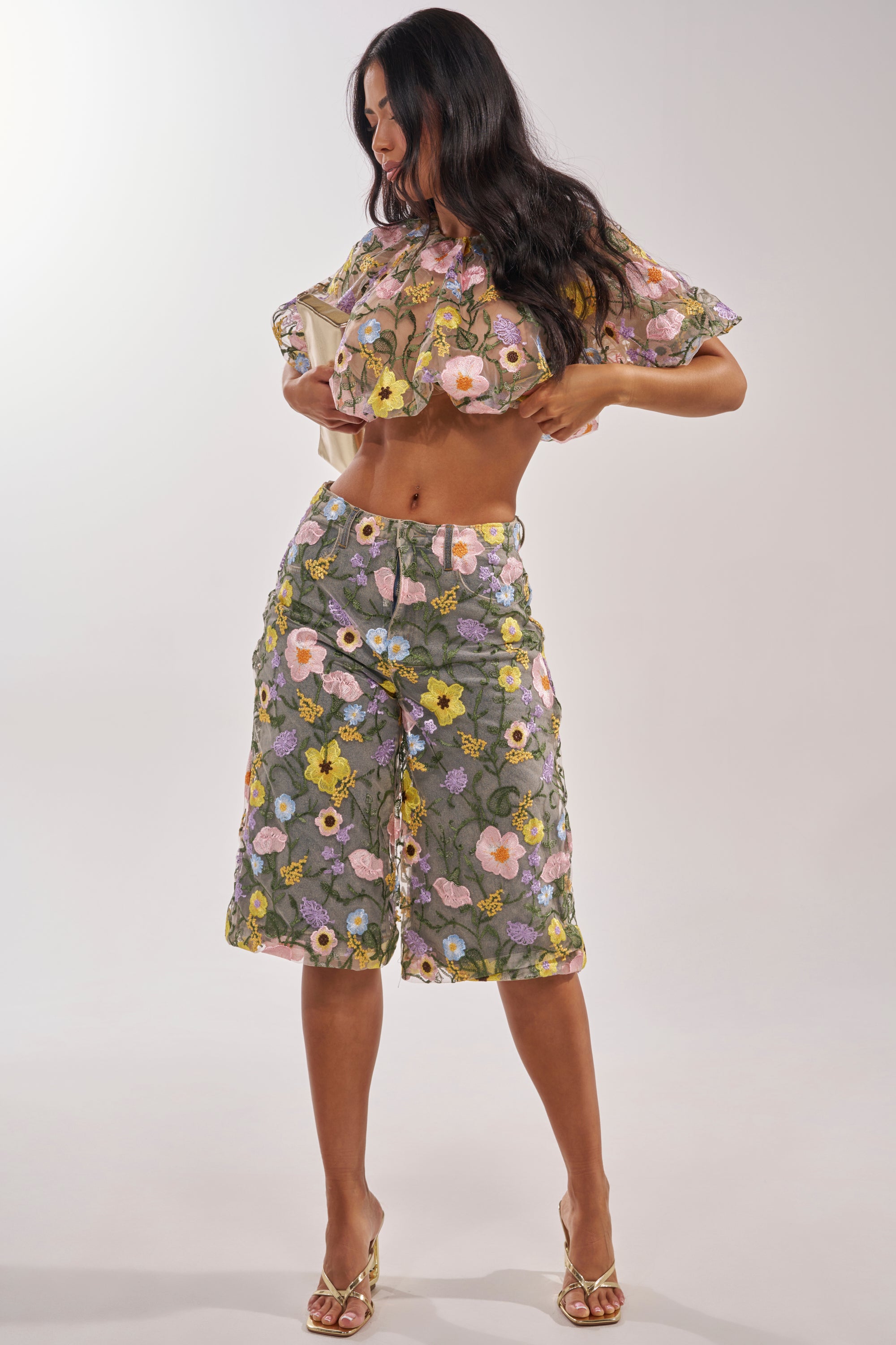 A woman poses confidently in GARDEN PARTY STRETCH DENIM WITH FLORAL TULLE BERMUDA SHORTS, paired with a cropped ruffled top and open-toed heels, standing against a plain light background. 7