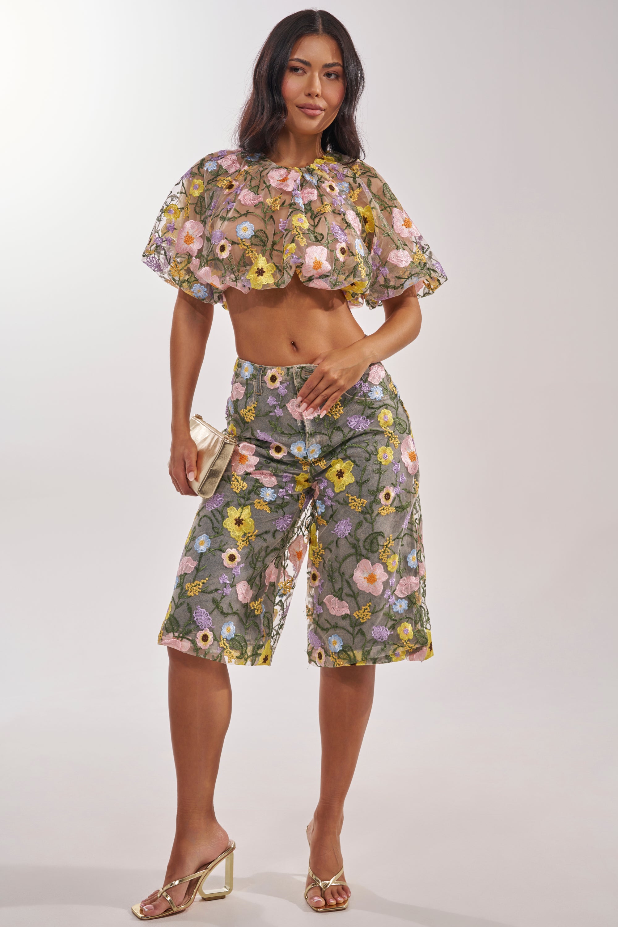A woman wears Garden Party Stretch Denim with Floral Tulle Bermuda Shorts, paired with a ruffled crop top. She holds a small clutch and gold heeled sandals, standing against a plain light background. 8
