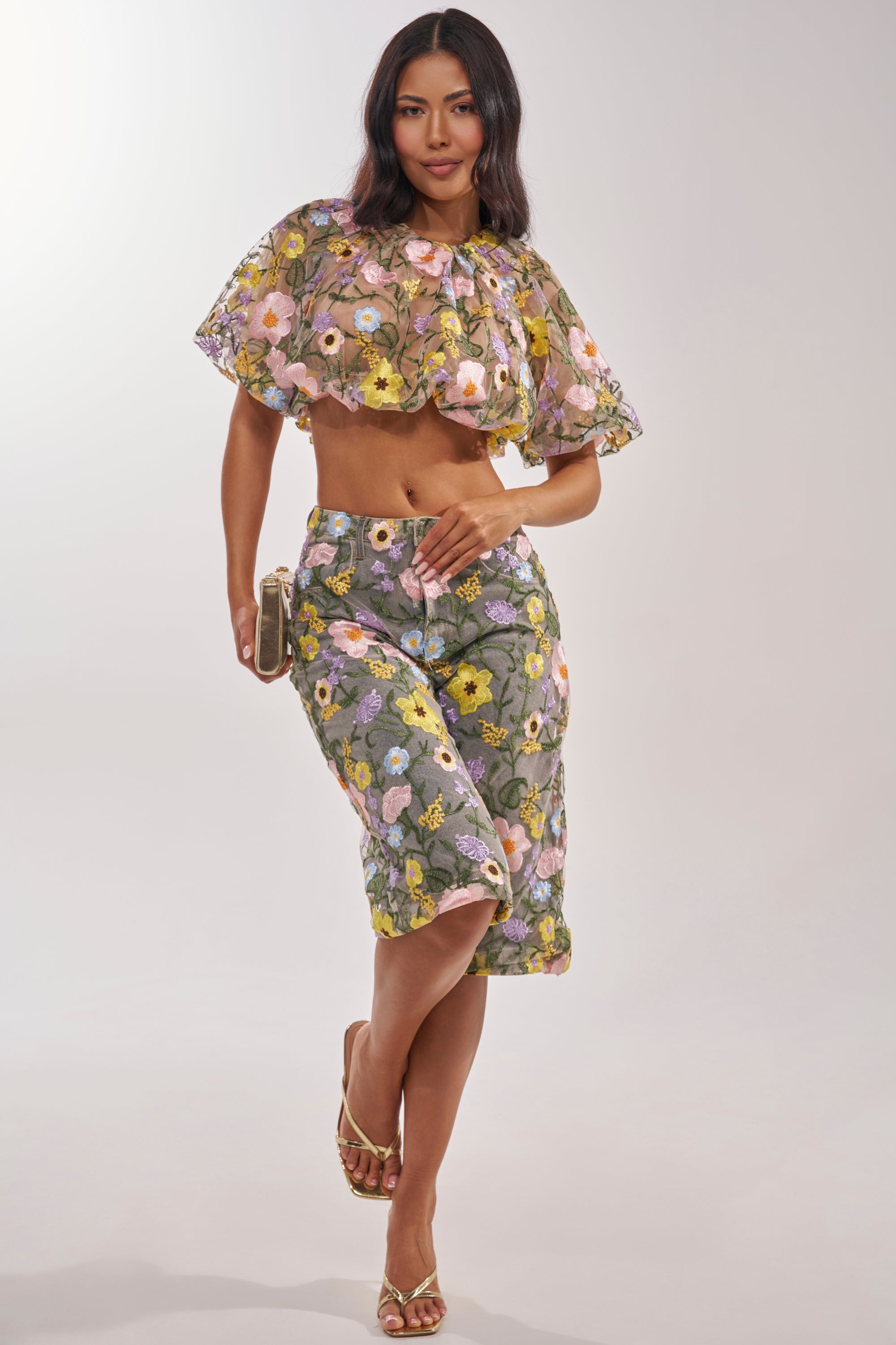 A woman poses in the GARDEN PARTY STRETCH DENIM WITH FLORAL TULLE BERMUDA SHORTS, paired with a cropped ruffled top. She holds a phone, wears gold sandals, and stands against a plain light gray background. 9