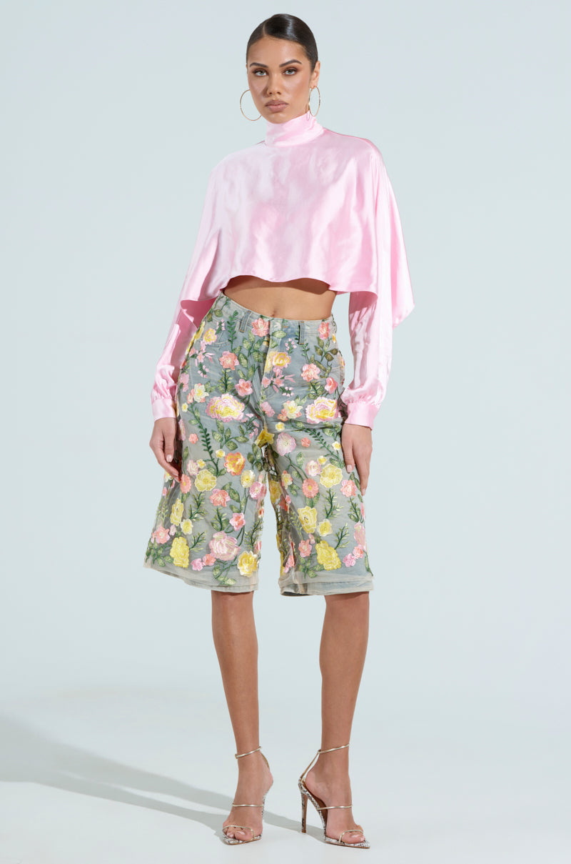 A person stands confidently in a pink, high-neck crop top and Garden Party Stretch Denim Bermuda Shorts with floral tulle. Silver hoop earrings and strappy high heels complete the look against a light gray backdrop.