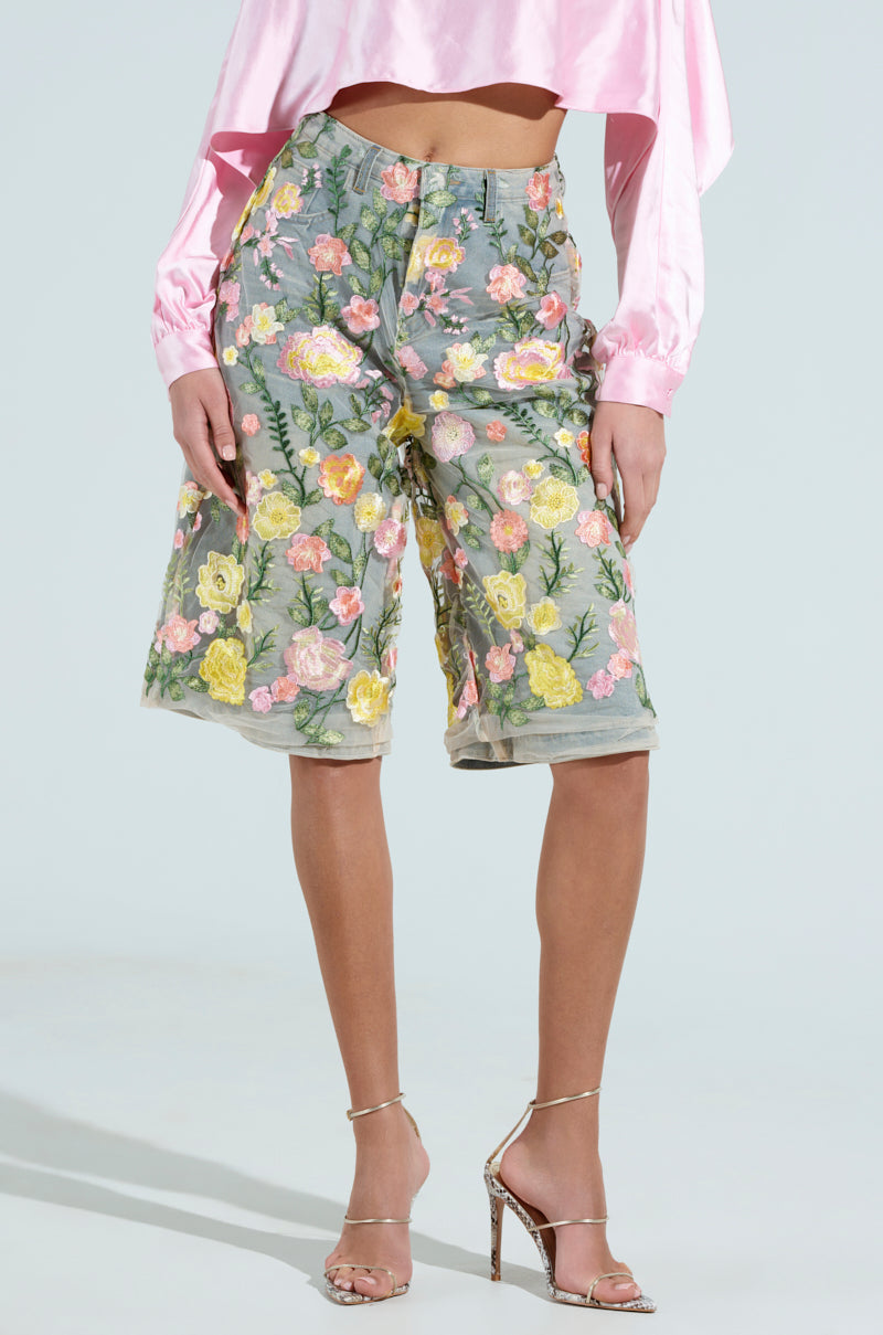Someone is wearing GARDEN PARTY STRETCH DENIM WITH FLORAL TULLE BERMUDA SHORTS in pink, yellow, and green over a gray base, paired with a pink cropped long-sleeve top and strappy high-heeled sandals.