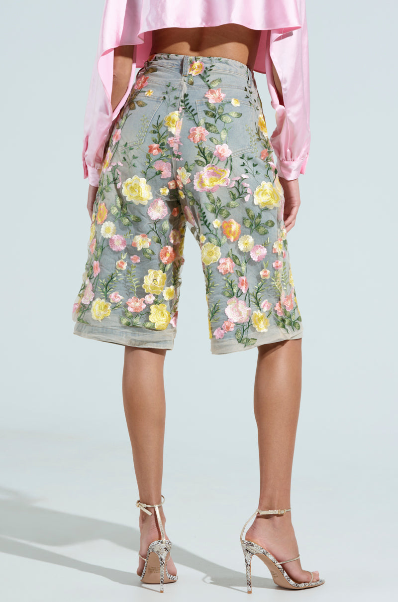 A person stands in a light, plain background, wearing a pink blouse with GARDEN PARTY STRETCH DENIM WITH FLORAL TULLE BERMUDA SHORTS in yellow and pink floral patterns, paired with high-heeled sandals.