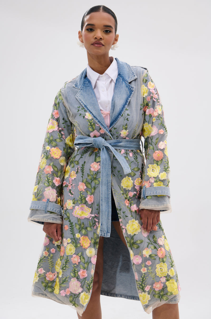 A woman poses confidently against a plain background, wearing the LIFE IN COLOR FLORAL DENIM TRENCH with a cinched silhouette and vibrant embroidery over a white shirt, her hands resting by her sides.