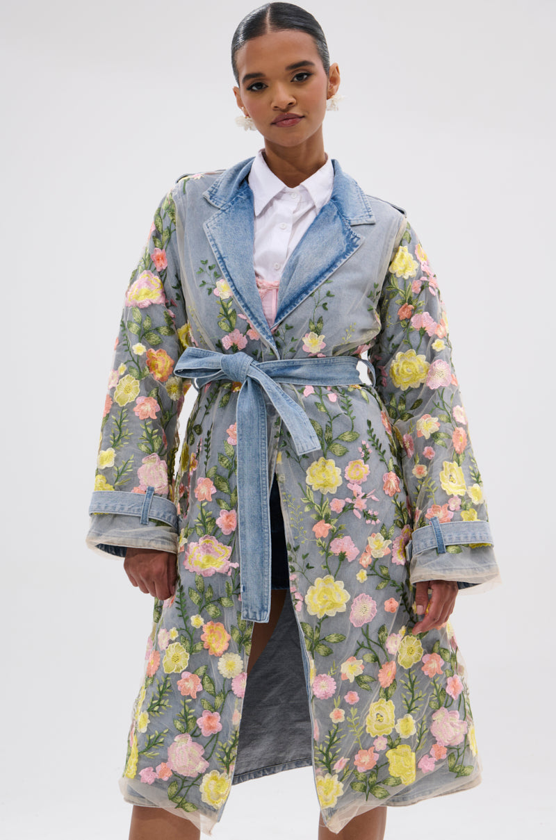 A woman wears the LIFE IN COLOR FLORAL DENIM TRENCH with yellow and pink embroidery over a white shirt, standing against a plain white background.