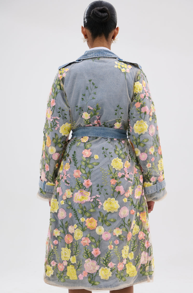 A person with dark hair in a bun is shown from the back, wearing the LIFE IN COLOR FLORAL DENIM TRENCH— a light blue trench adorned with colorful yellow and pink floral embroidery, set against a plain white background.