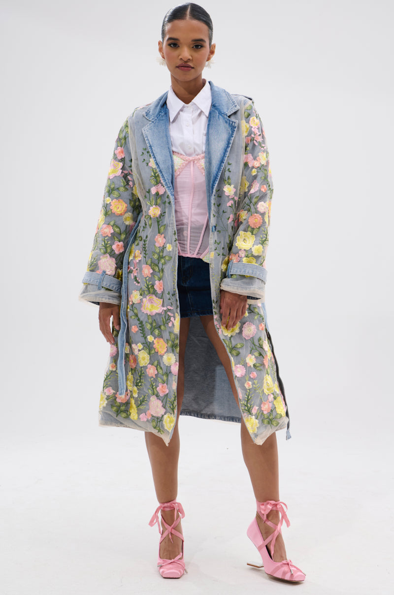 A woman stands confidently against a white background, wearing the LIFE IN COLOR FLORAL DENIM TRENCH over a denim jacket, white shirt, and shorts, paired with pink lace-up shoes and her hair tied back.