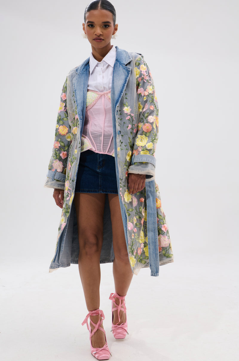 A woman stands confidently against a white background wearing the LIFE IN COLOR FLORAL DENIM TRENCH over a white shirt, pink corset, denim mini skirt, and pink lace-up heels, creating a striking cinched silhouette.