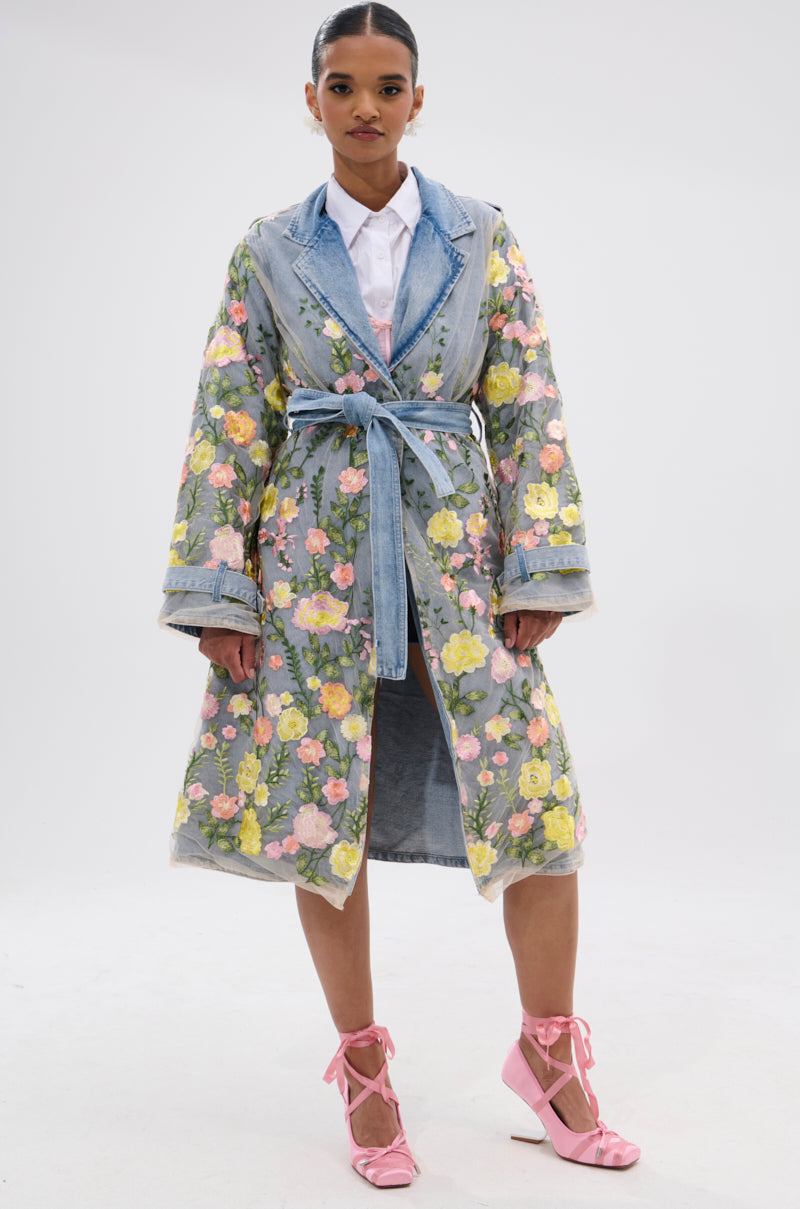 A woman faces forward against a white background, wearing the LIFE IN COLOR FLORAL DENIM TRENCH with sheer floral embroidery over a white shirt and pink lace-up heels.