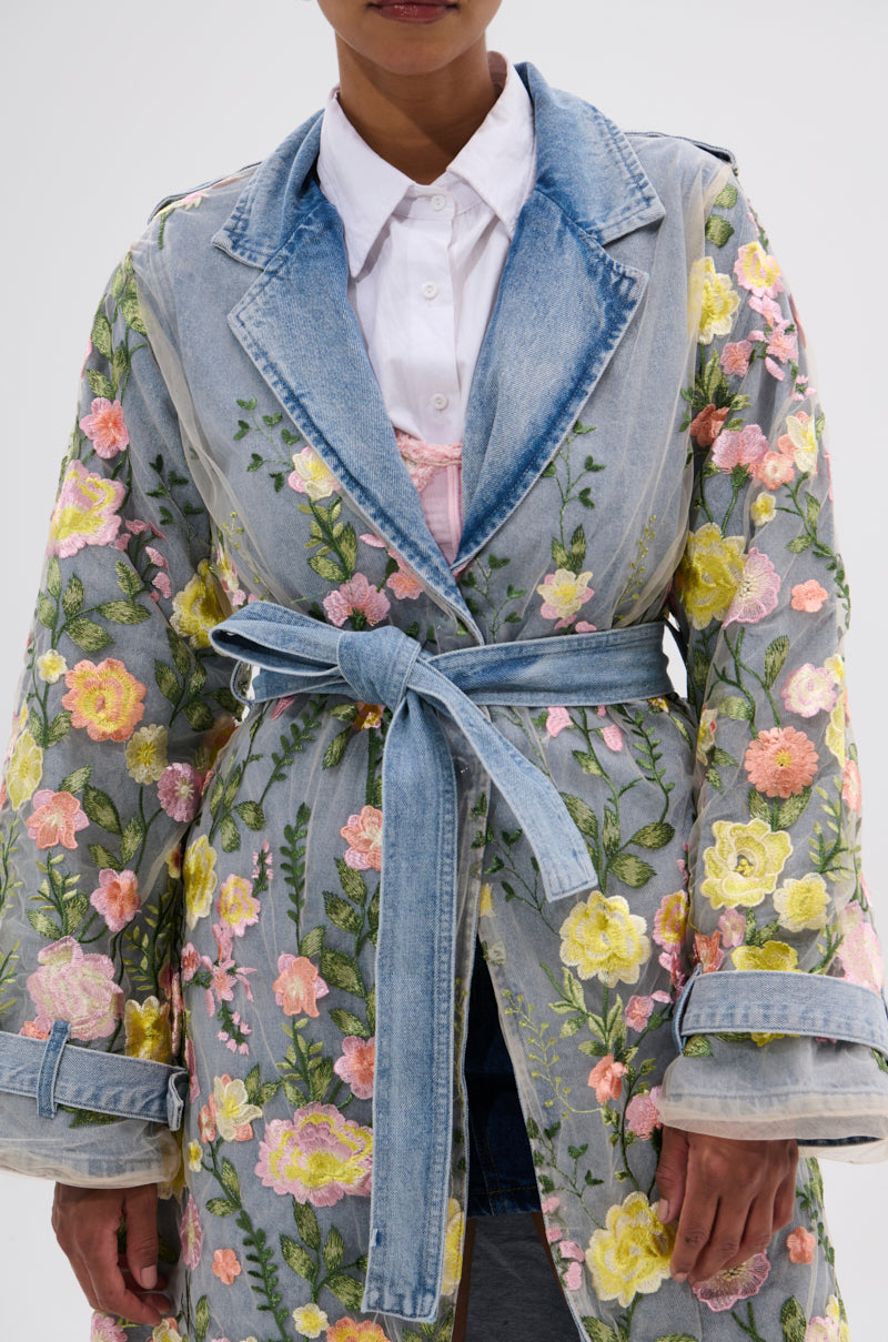 A person wears the LIFE IN COLOR FLORAL DENIM TRENCH—a light blue, belted coat with a cinched silhouette and colorful embroidery—over a white collared shirt in an image cropped at the shoulders and knees.