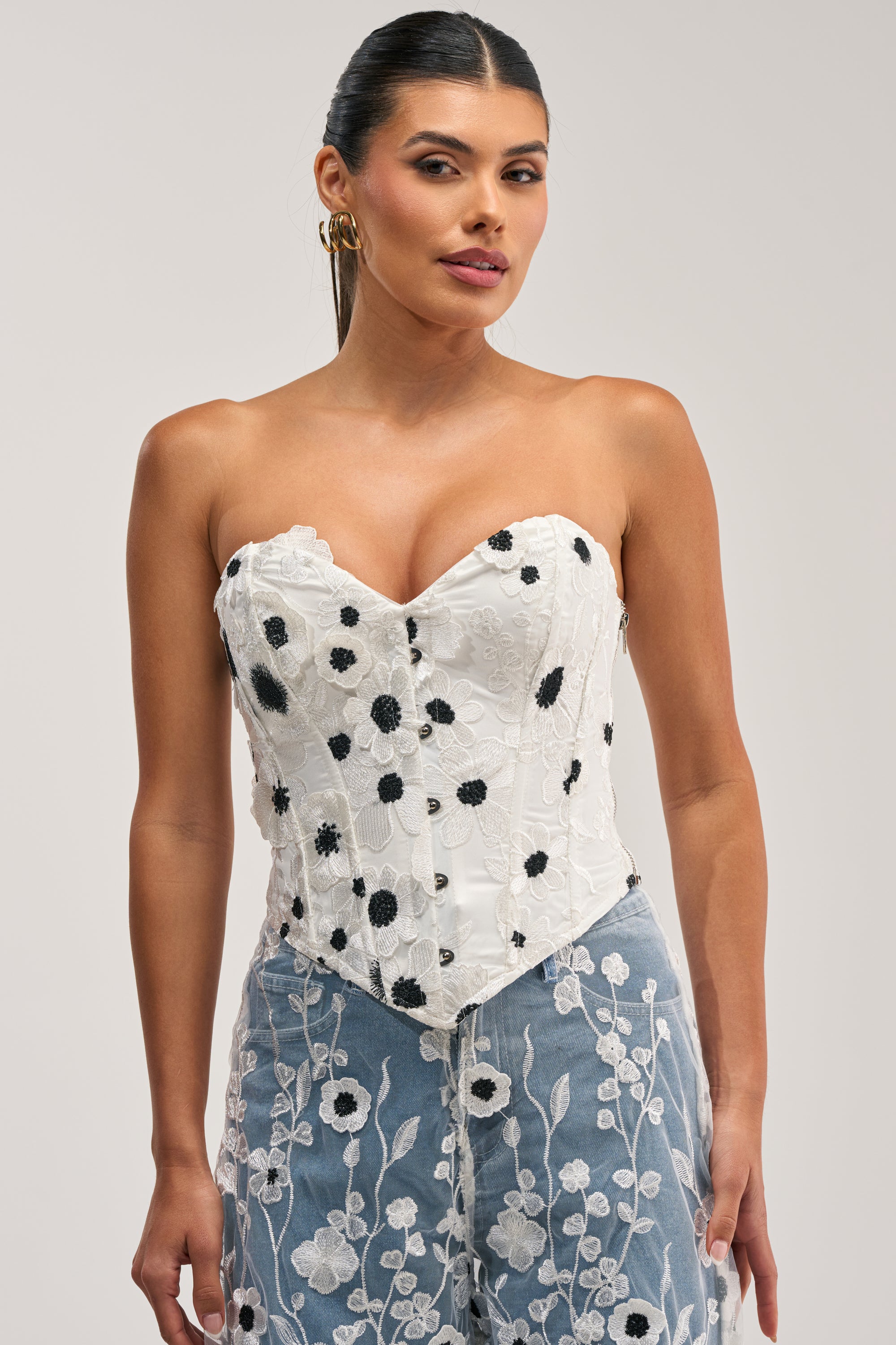 A woman models the MIXED FLORALS CORSET, a strapless white piece featuring black floral patterns and a strapless neckline, styled with high-waisted sheer pants adorned with white floral embroidery, posed against a plain light background.