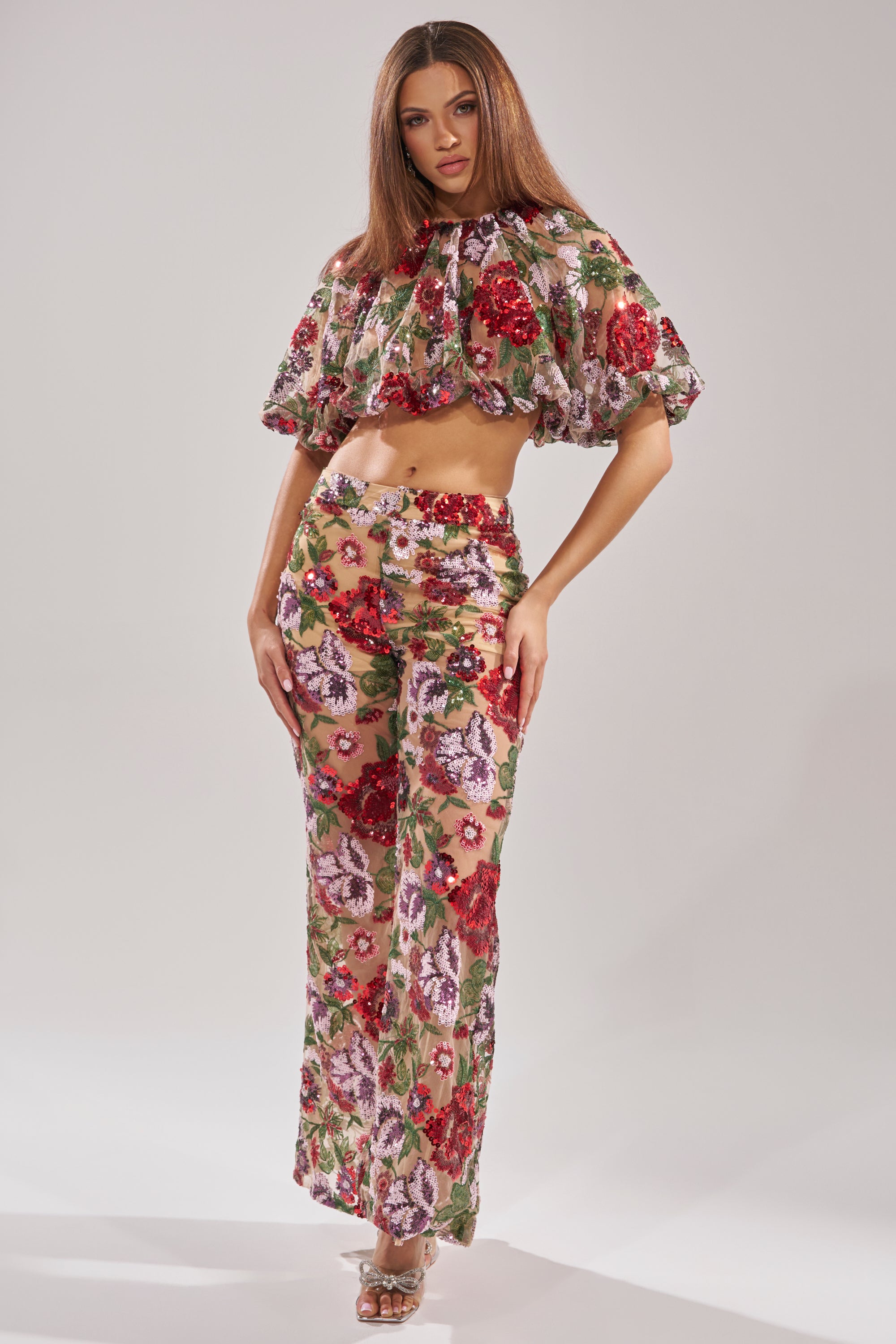 A woman poses in ENCHANTED GARDEN FLORAL EMBROIDERED TULLE PANTS, paired with a matching sheer ruffled crop top in red, pink, and green florals. Her long hair flows freely and she finishes the look with clear high-heeled shoes.