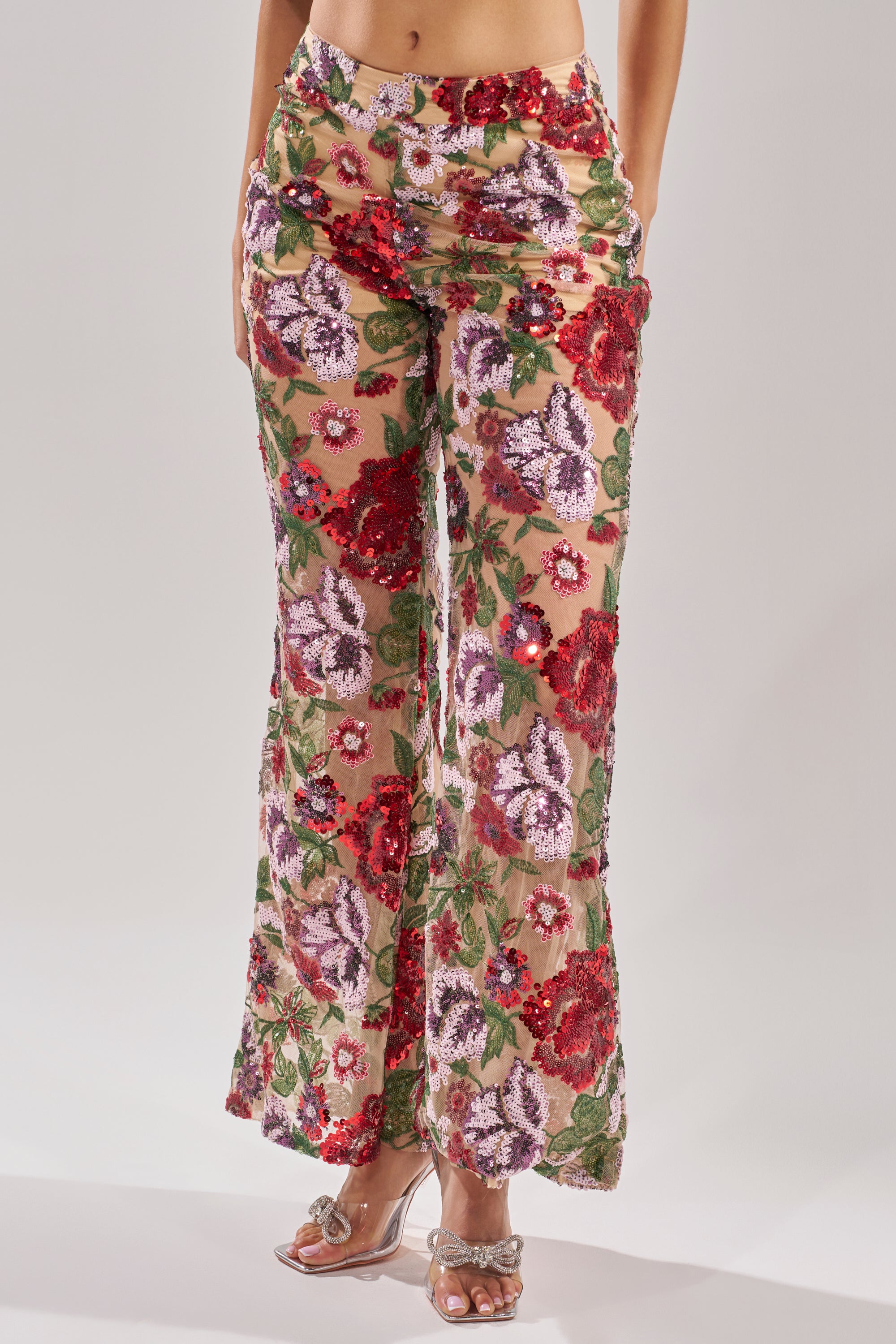 A person wears the ENCHANTED GARDEN FLORAL EMBROIDERED TULLE PANT, high-rise flares in sheer mesh with red, purple, and green floral embroidery, styled with clear heeled sandals. Photo is cropped midriff to feet.
