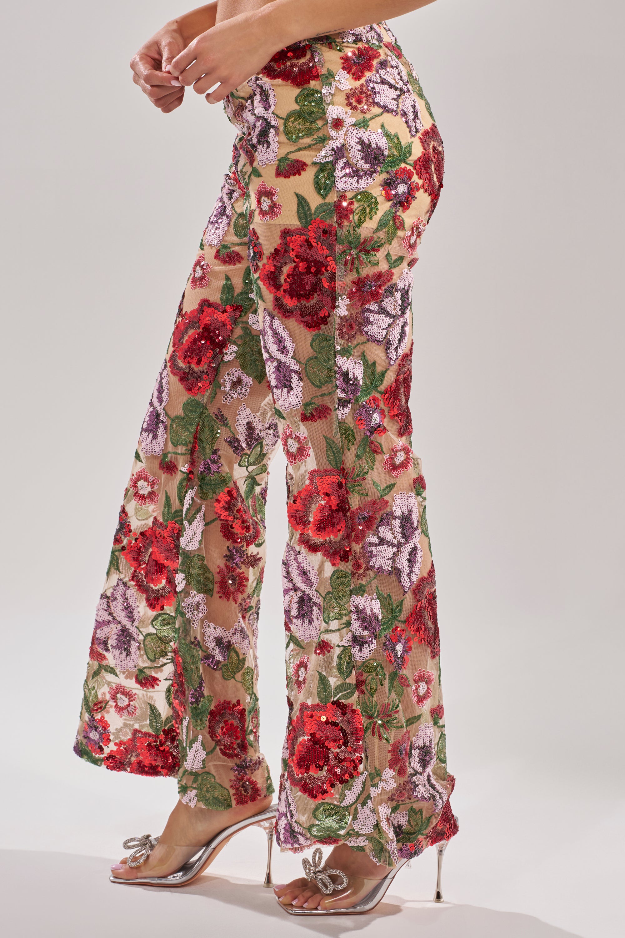 A person wears the ENCHANTED GARDEN FLORAL EMBROIDERED TULLE PANT, featuring high-rise flared sheer mesh with red, purple, and green floral embroidery, styled with transparent high heels with bow accents against a plain background.