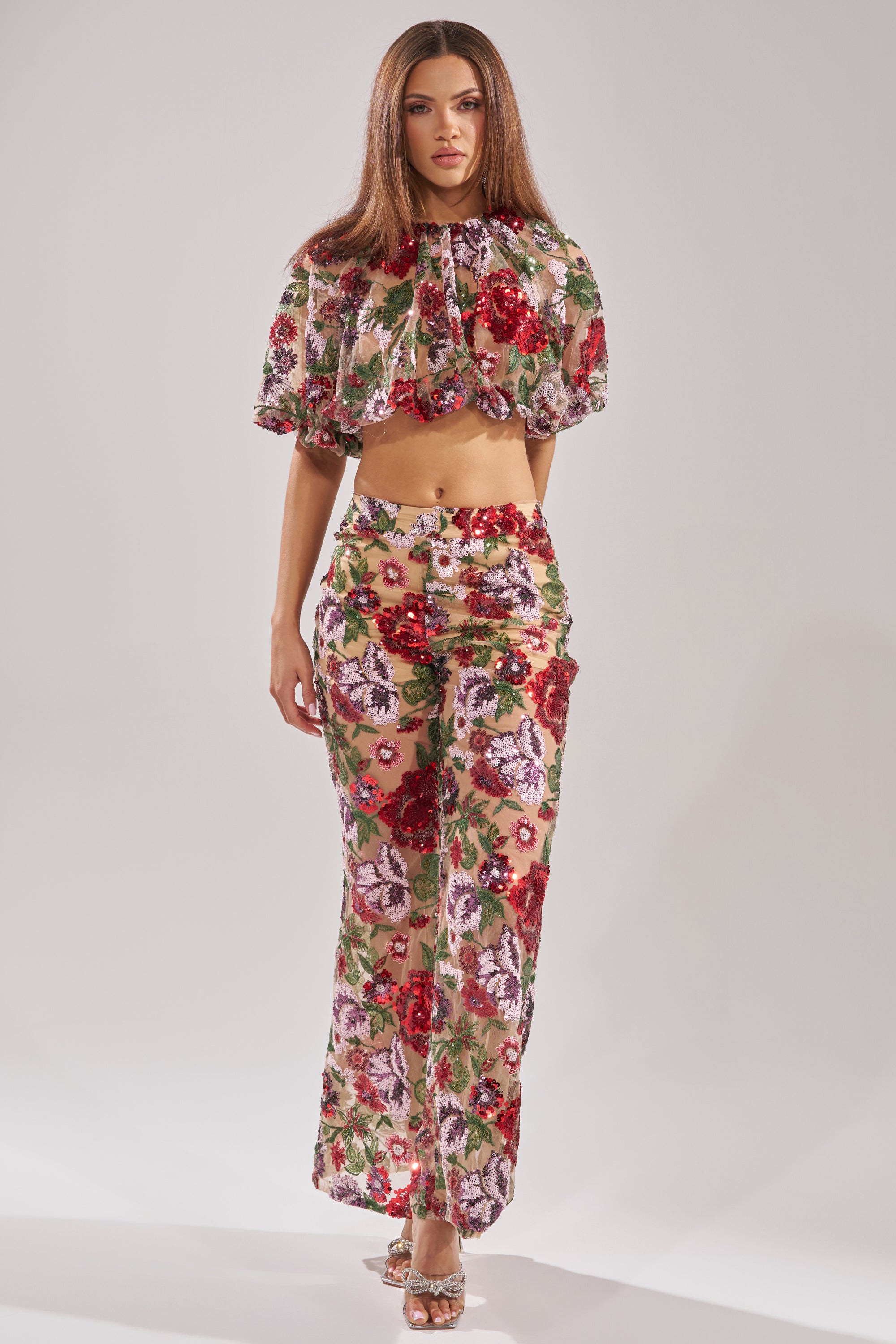 A woman models the ENCHANTED GARDEN FLORAL EMBROIDERED TULLE PANT with a matching cropped, puff-sleeve top featuring floral sequin accents. She pairs the sheer set with heeled sandals and wears her straight hair loose against a plain background.