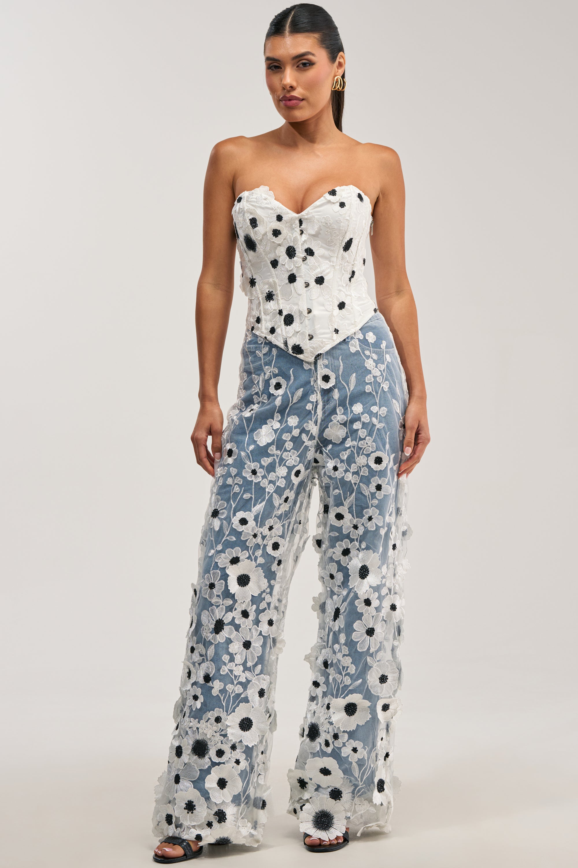 A woman stands against a plain background wearing the MIXED FLORALS RELAXED DENIM—high-waisted denim pants adorned with white and black floral embroidery and a delicate tulle overlay. She pairs them with a strapless white corset featuring black accents.