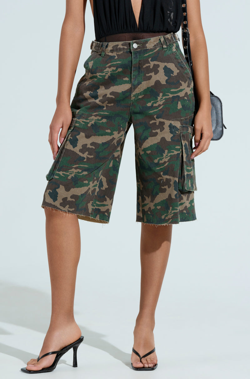 Wearing a black sleeveless top and "Down and Dirty Camo Bermuda Short," they pose against a light background, holding a black bag. The look is completed with black high-heeled sandals, effortlessly blending style with function. 1