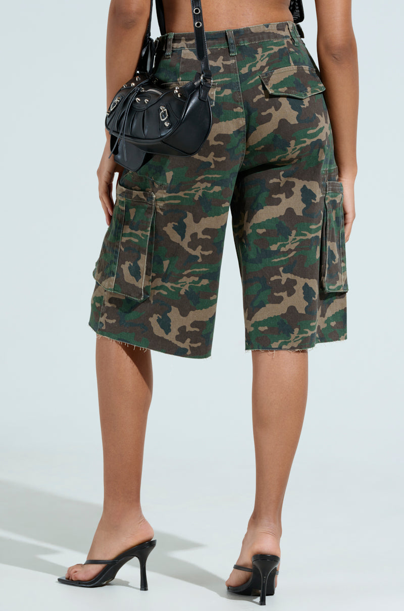 Wearing DOWN AND DIRTY CAMO BERMUDA SHORT and black high-heeled sandals, a person stands with their back to the camera, holding a small black handbag with a shoulder strap against a light-colored backdrop. 3