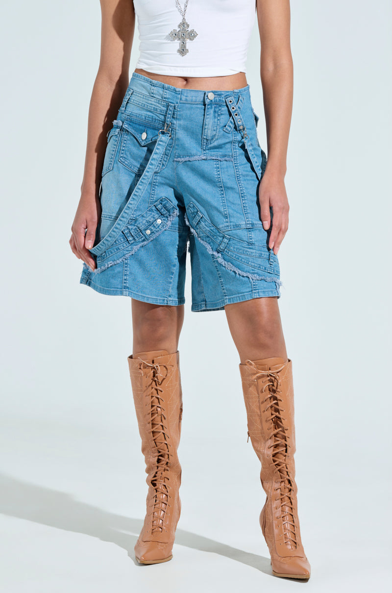 Someone is wearing a white crop top with GRAB ME CLOSER DENIM BERMUDA SHORT featuring unique patchwork and straps, paired with tan knee-high lace-up boots. A silver necklace accessorizes the look against a plain white background.