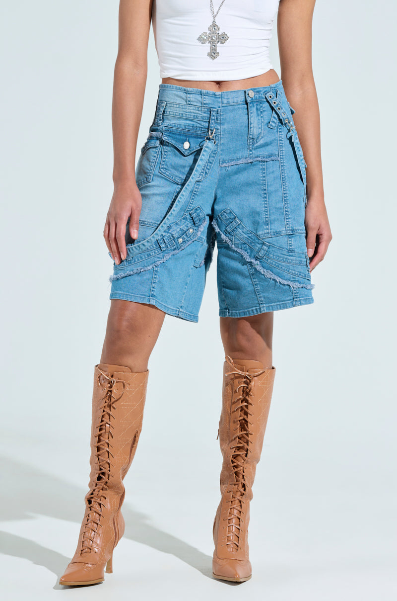 A person in a white sleeveless top, a long silver cross necklace, GRAB ME CLOSER DENIM BERMUDA SHORT with a low-rise fit and multiple pockets and straps, and tan lace-up knee-high boots stands against a plain background.