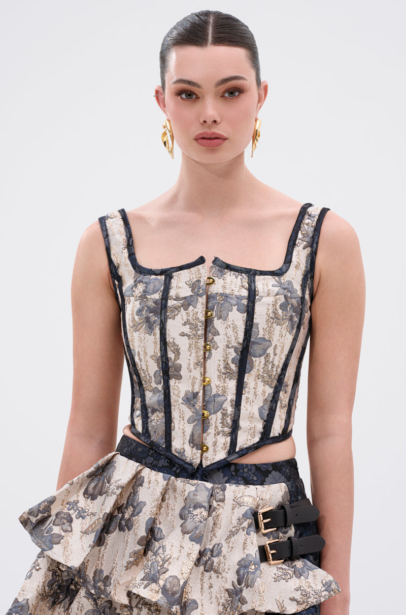 A woman with slicked-back hair wears the WON'T STOP MOVING BROCADE CORSET, featuring metallic floral brocade, gold buttons, black trim, and pairs it with a matching skirt. She accessorizes with bold gold earrings. 0