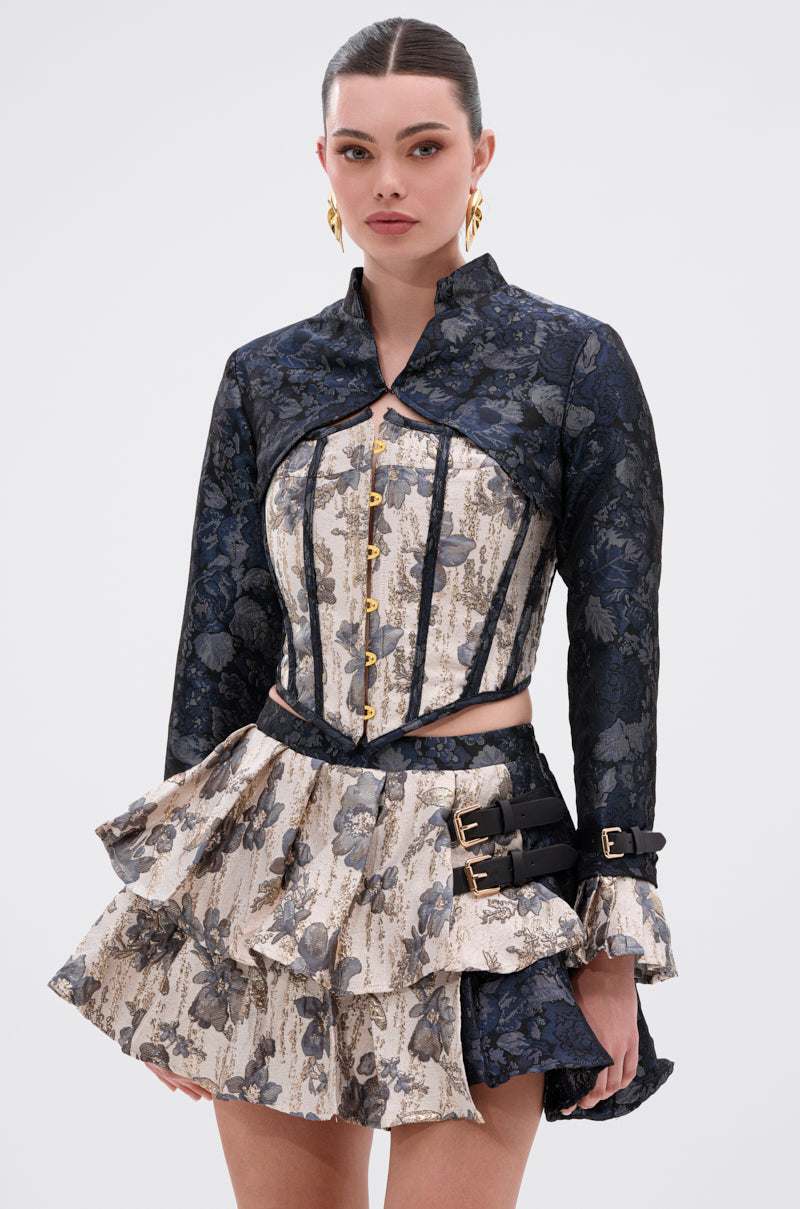 A woman stands against a plain light background wearing the WON'T STOP MOVING BROCADE CORSET dress, featuring a ruffled floral print, brocade corset bodice, long sleeves, lace-up back, and buckle details on the skirt. 1