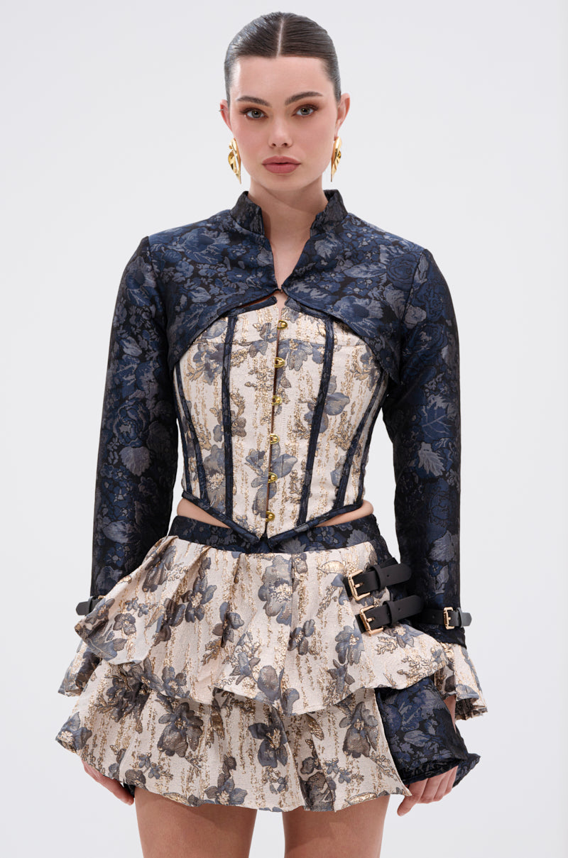 A woman wears the WON'T STOP MOVING BROCADE CORSET with a metallic floral design and lace-up back, styled with a dark jacket, layered ruffled skirt, slicked-back hair, and gold hoop earrings against a plain background. 2