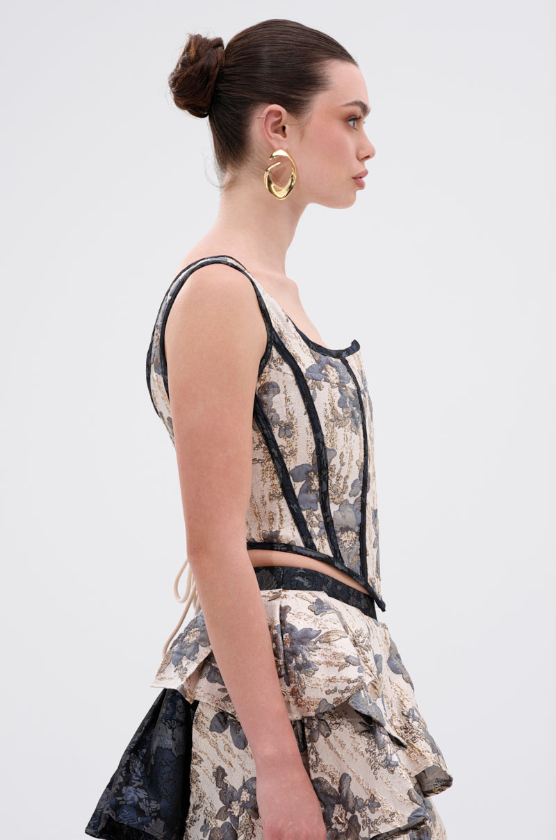 A woman with her brown hair in a bun wears large gold hoop earrings and the WON'T STOP MOVING BROCADE CORSET peplum dress in beige and blue with a lace-up back, standing in profile against a plain light background. 3