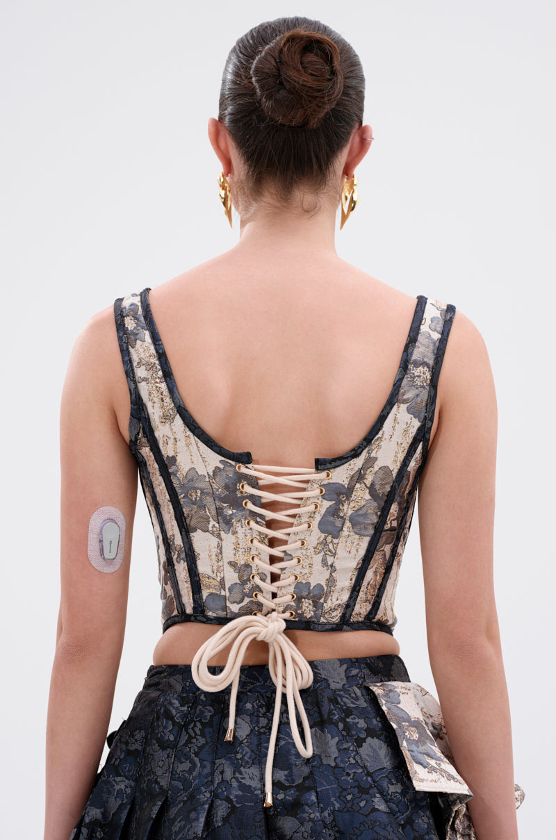 A person with dark hair in a bun wears the WON'T STOP MOVING BROCADE CORSET and skirt, featuring a lace-up back. A medical patch is visible on their upper left arm. Photo taken from behind against a plain background. 4