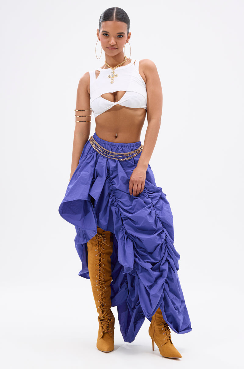 A woman poses confidently in an AKIRA Label white cut-out crop top, the NOT ABOUT IT RUCHED MIDI SKIRT with an asymmetrical hem and gold chain detail, tan lace-up knee-high boots, large hoop earrings, and gold arm jewelry. 0