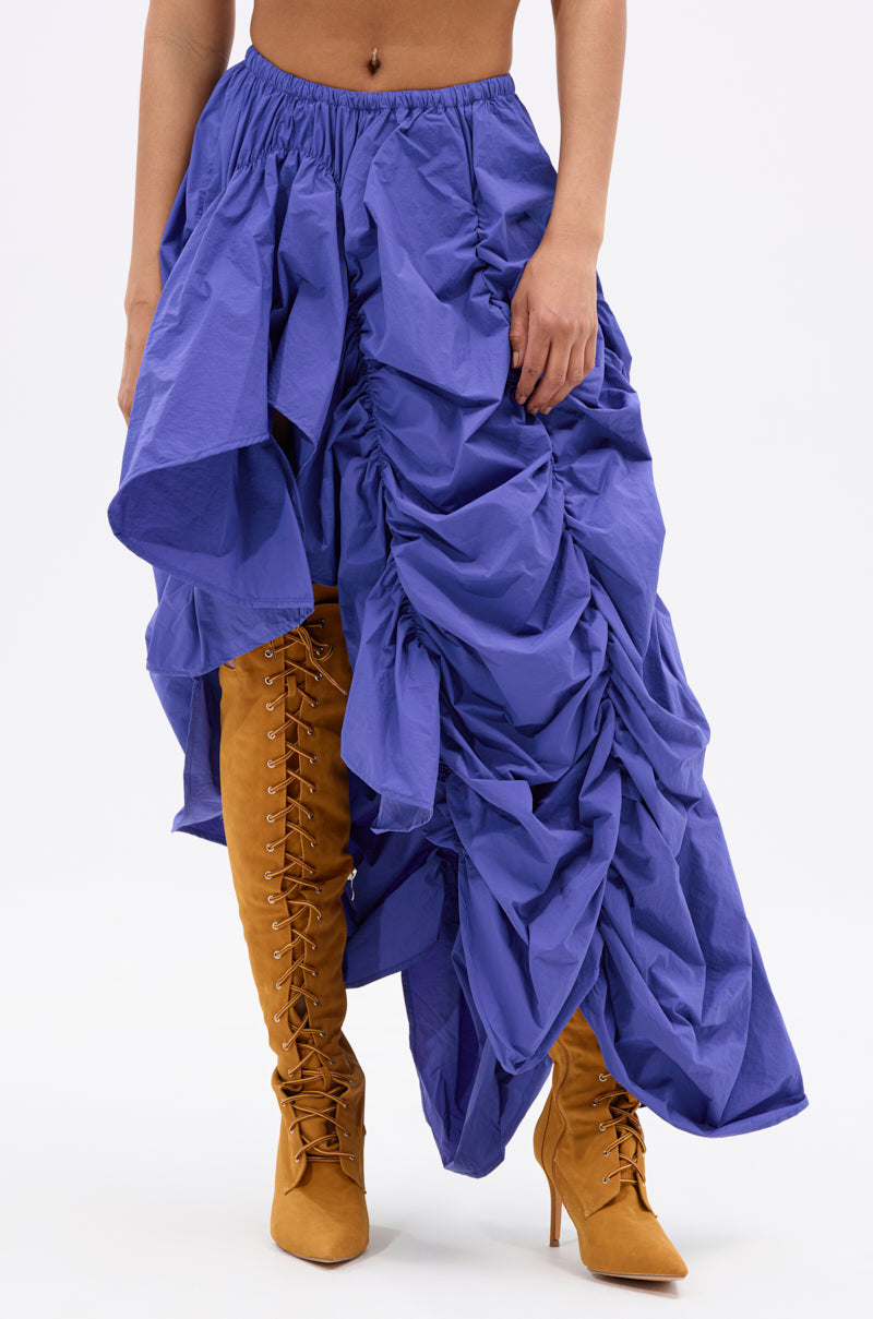 A person models the NOT ABOUT IT RUCHED MIDI SKIRT in bright blue with an asymmetrical hem, styled with high-heeled tan lace-up boots. The photo is cropped at the waist and set against a plain white background. 1