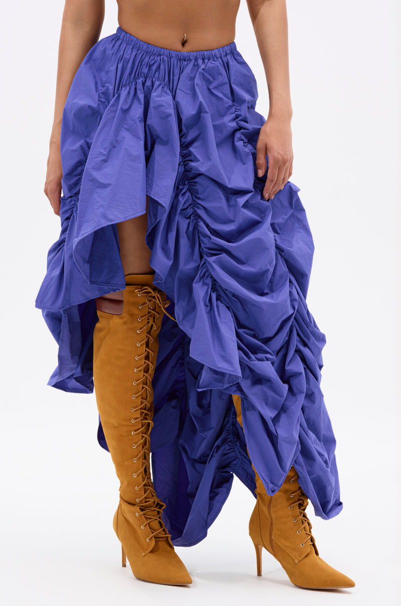 A person models the NOT ABOUT IT RUCHED MIDI SKIRT in bright blue with an asymmetrical hem, styled with tan knee-high, lace-up pointed-toe boots. Only their lower torso and legs appear against a plain white background. 2