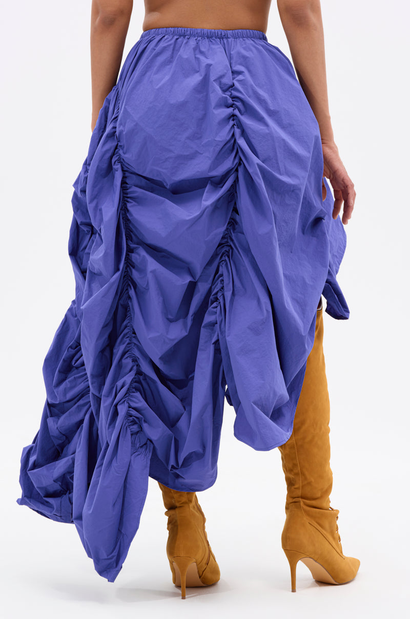 A person in the NOT ABOUT IT RUCHED MIDI SKIRT, bright blue with an asymmetrical hem, and tall mustard-yellow heeled boots, stands with their back to the camera against a plain white background. 5