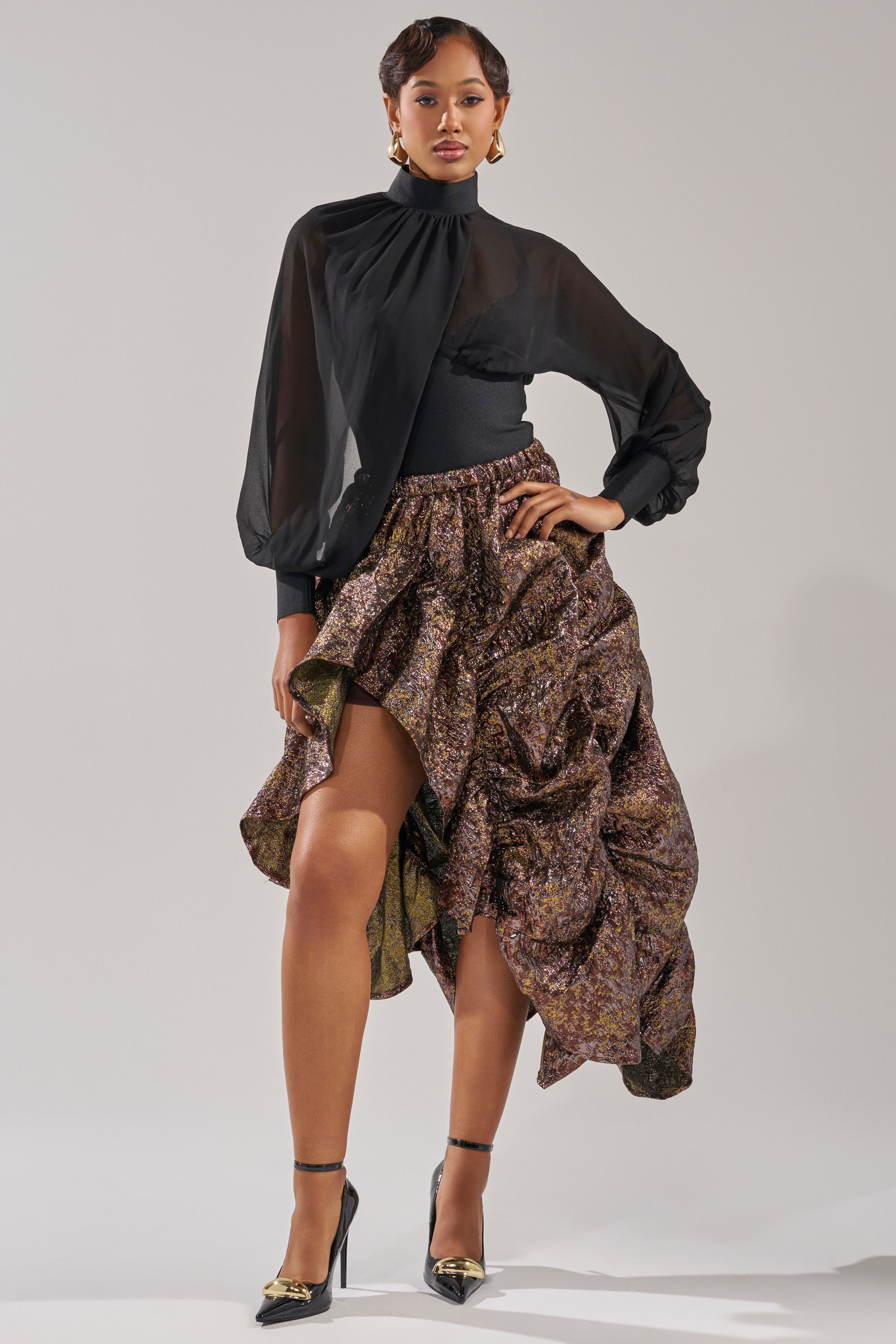 A woman poses confidently in the CRAZY FOR YOU BROCADE RUCHED MIDI SKIRT, styled with a black sheer blouse, black and gold heels, and statement earrings against a plain gray background. 0