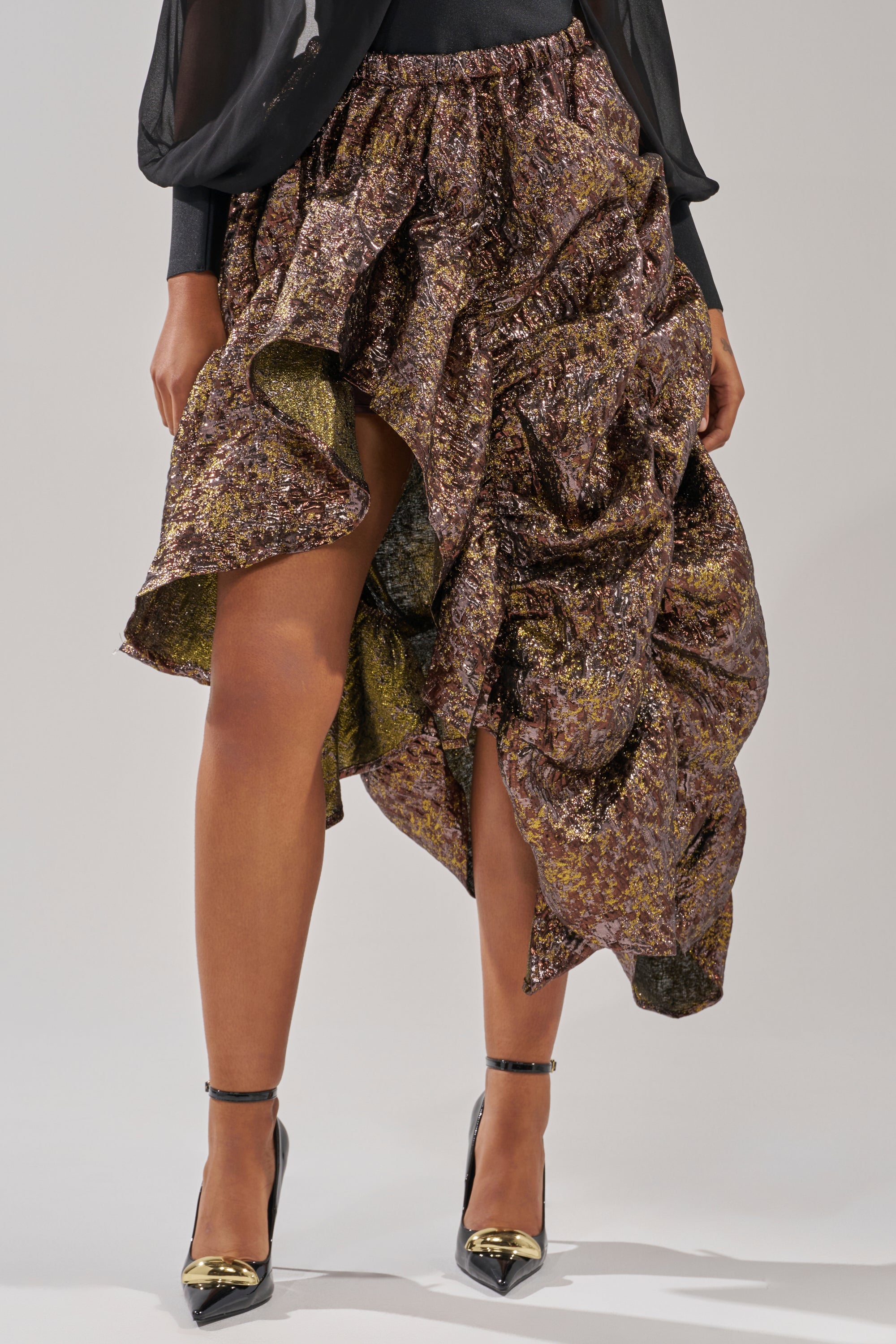 Wearing the CRAZY FOR YOU BROCADE RUCHED MIDI SKIRT, styled with black heels featuring gold details, showcases bold textures and dramatic draping with its metallic sheen and unique ruching. 2