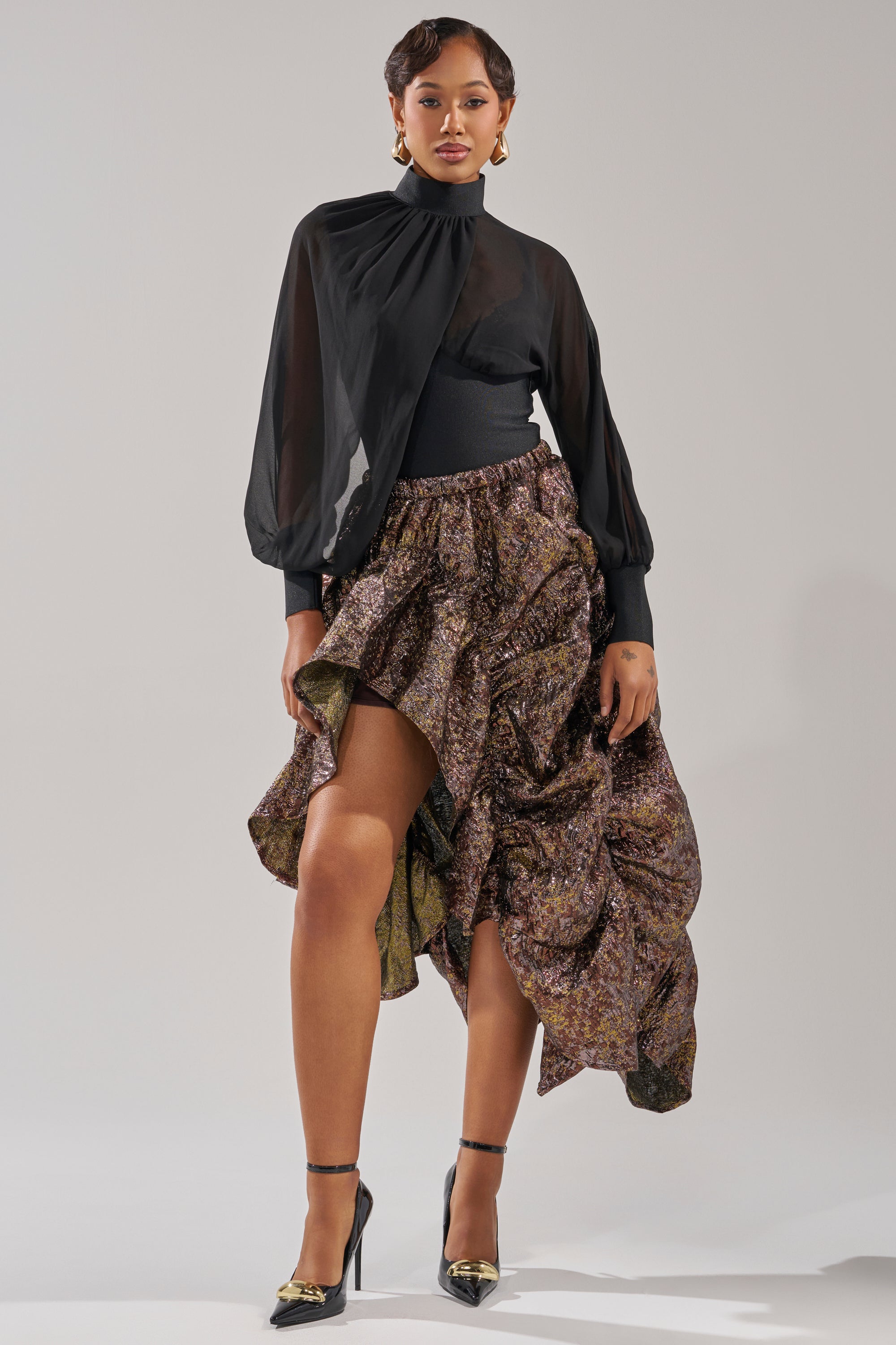 A woman poses in a black sheer long-sleeve blouse paired with the CRAZY FOR YOU BROCADE RUCHED MIDI SKIRT, styled with black heels featuring gold details and statement earrings, all set against a plain light background. 6