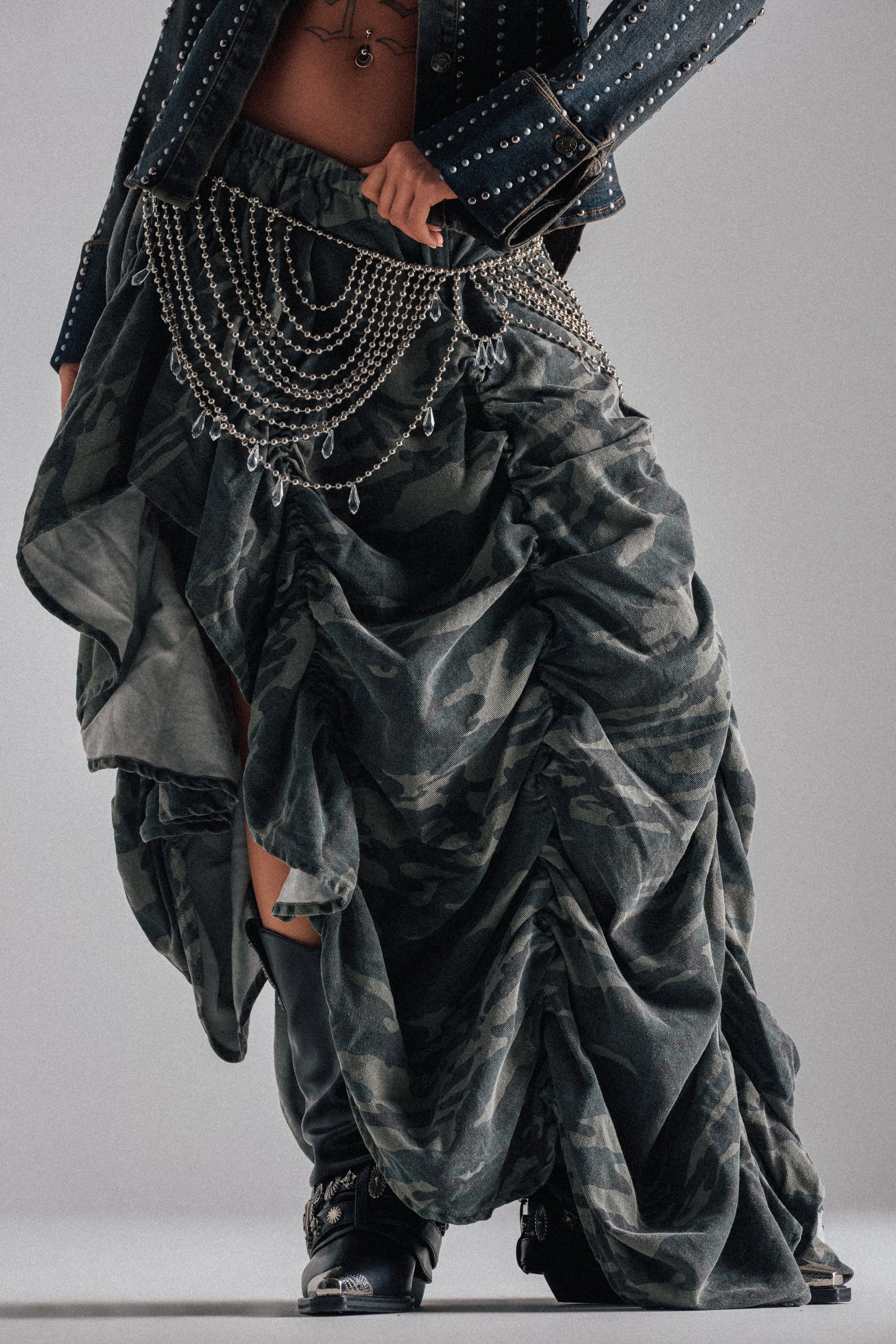Person wearing the LETS CHANGE IT UP RUCHED MIDI SKIRT IN CAMO with a metal chain belt, dark studded jacket, and black boots. The chest-down crop highlights statement streetwear details and bold style elements.