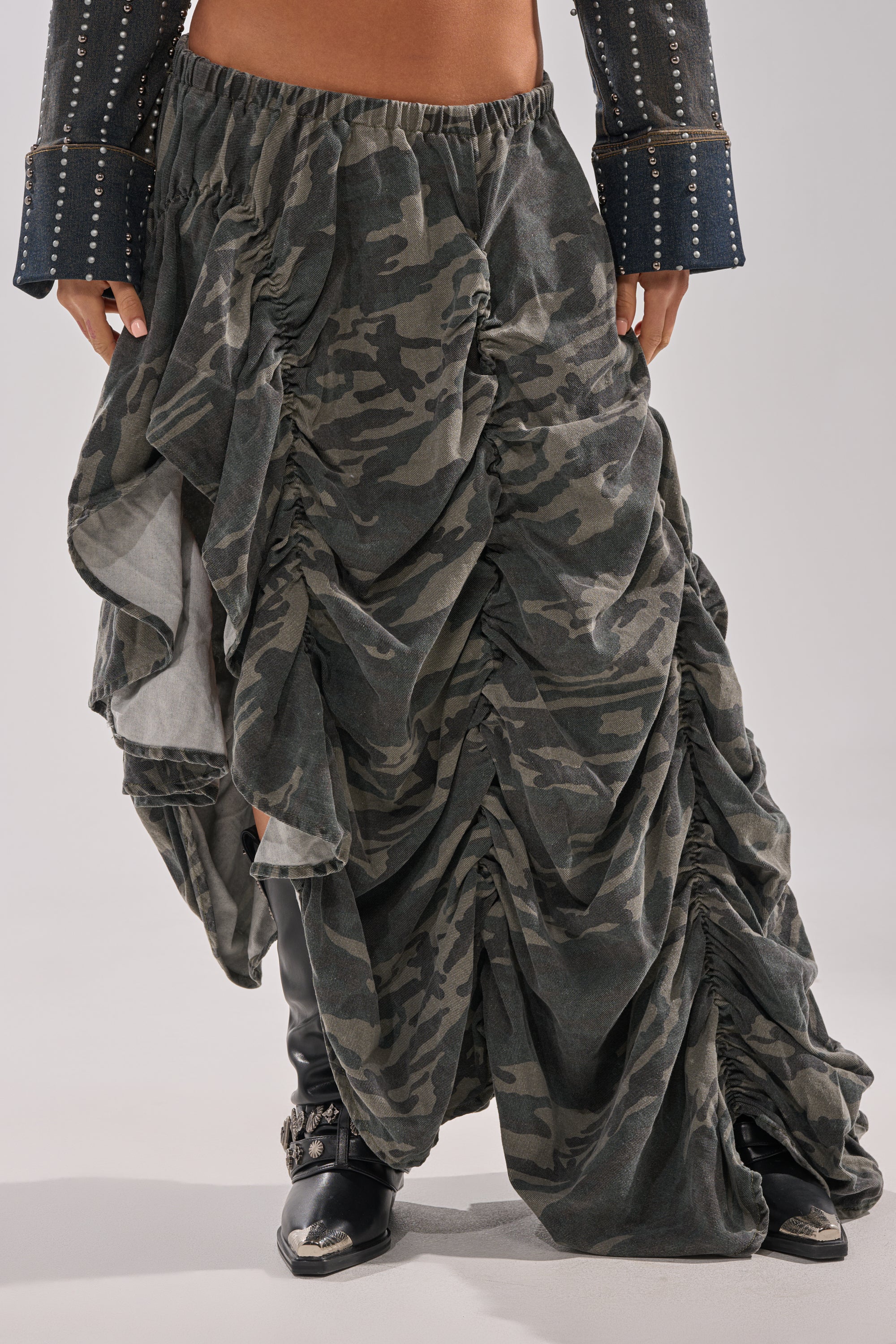 Wearing the LETS CHANGE IT UP RUCHED MIDI SKIRT IN CAMO with dramatic draping and ruffles, paired with black knee-high boots and a dark studded-cuff jacket for an edgy streetwear vibe.