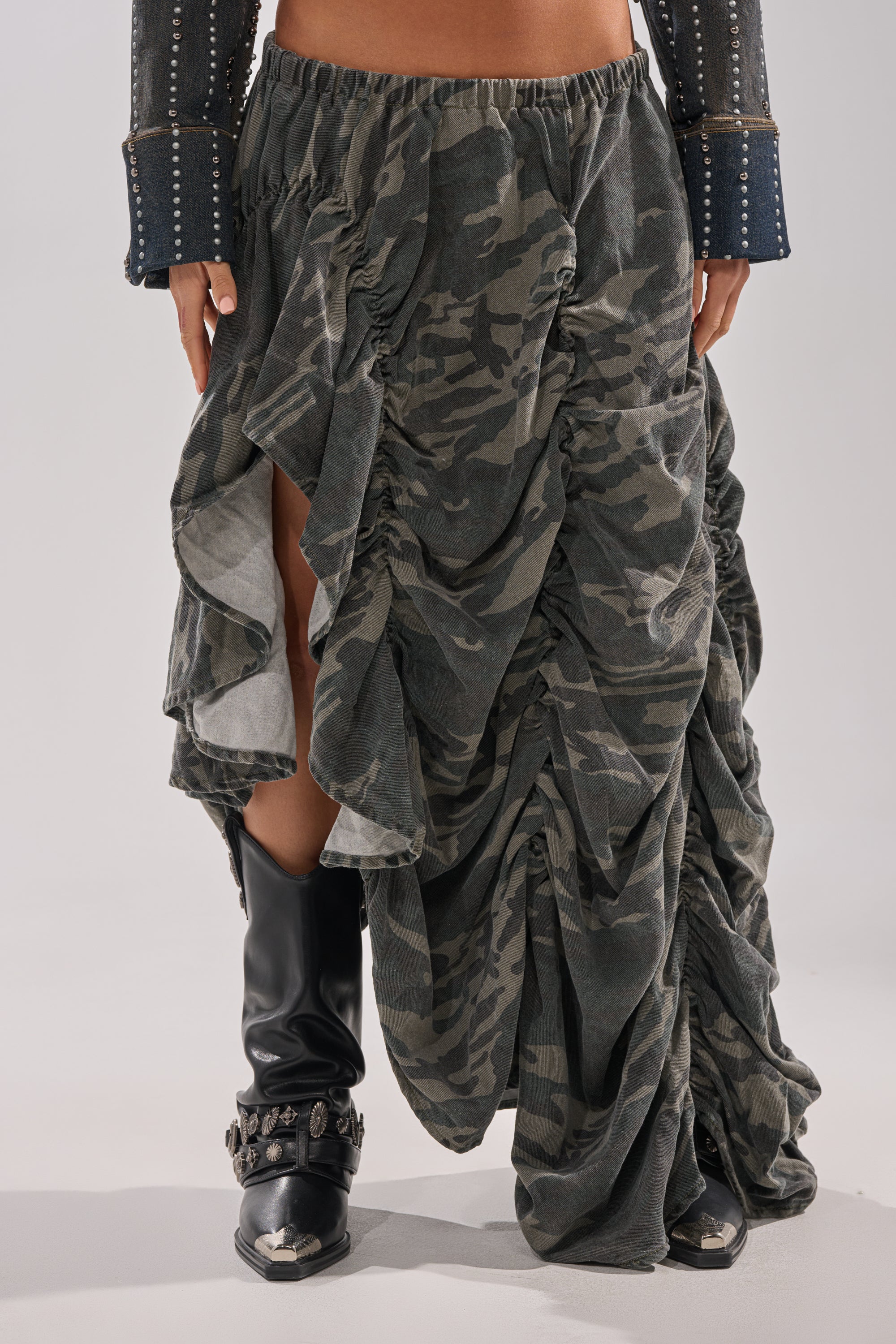 A person is seen in the LETS CHANGE IT UP RUCHED MIDI SKIRT IN CAMO, styled with black studded cowboy boots and a jacket featuring studded sleeves. The photo is cropped to display only the lower half of the body.