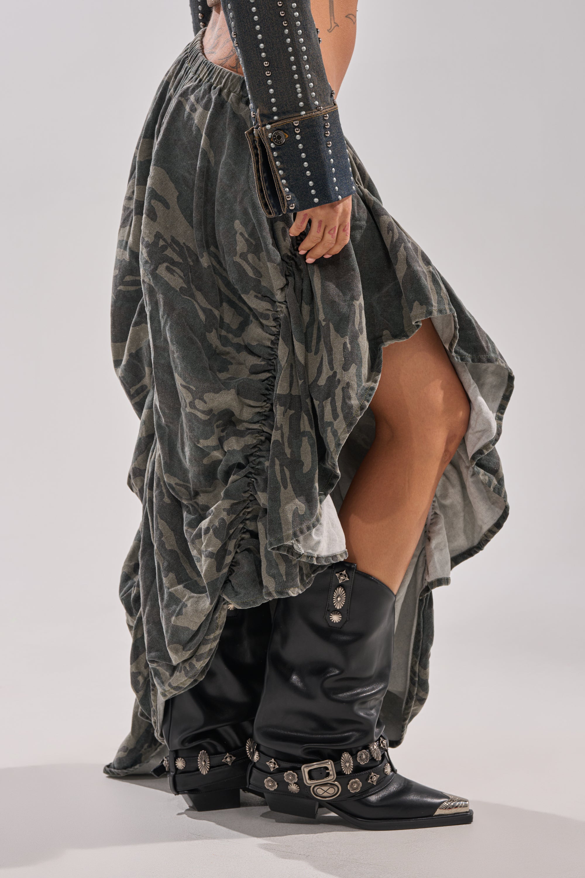 Wearing the LETS CHANGE IT UP RUCHED MIDI SKIRT IN CAMO with a high slit, paired with black studded cowboy boots and a matching arm accessory, creates a bold streetwear statement against a white background.