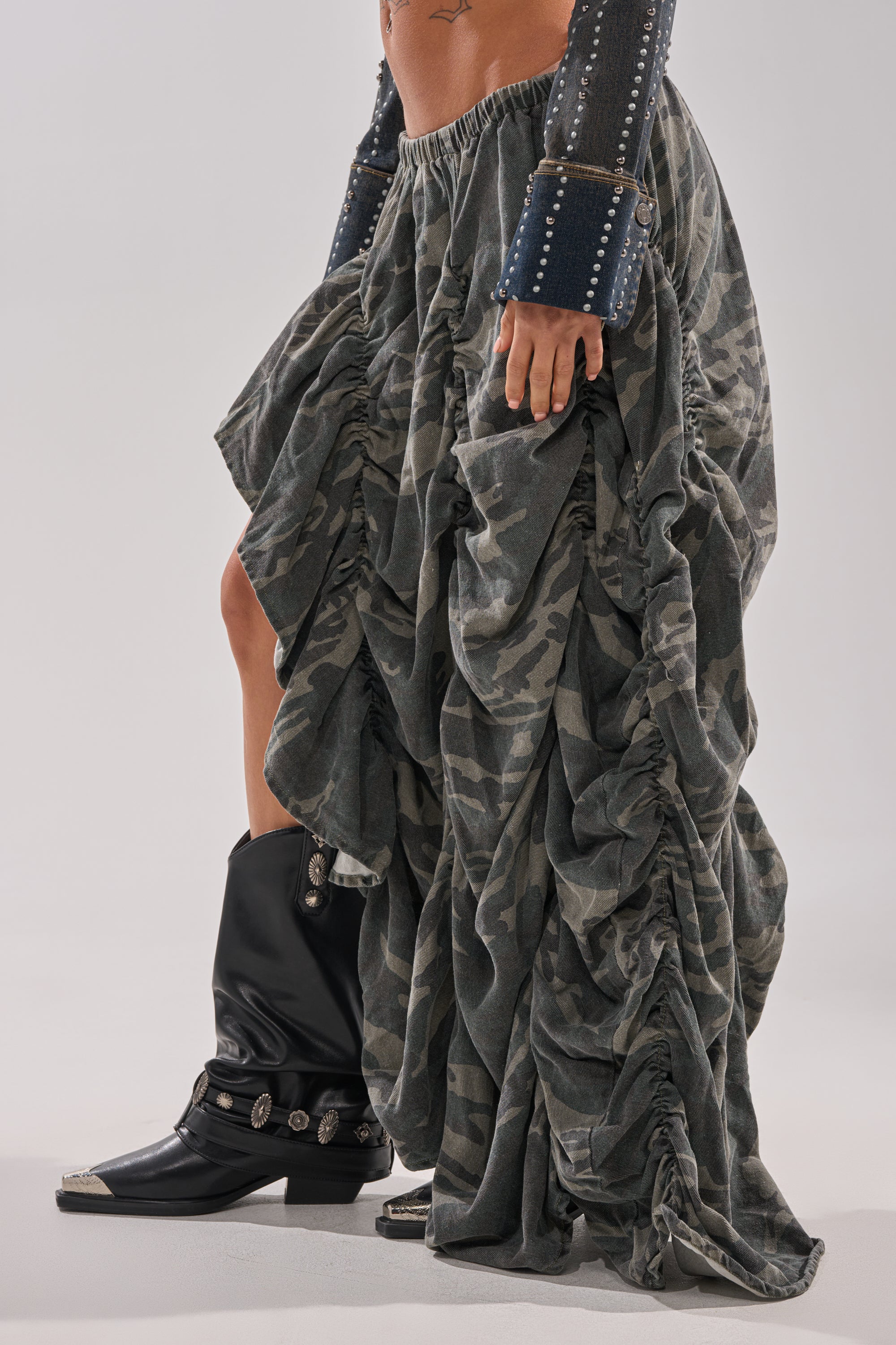 Wearing the LETS CHANGE IT UP RUCHED MIDI SKIRT IN CAMO with an asymmetrical draped silhouette, a studded denim jacket, and black western-style boots, a person poses against a plain background in a bold streetwear look.